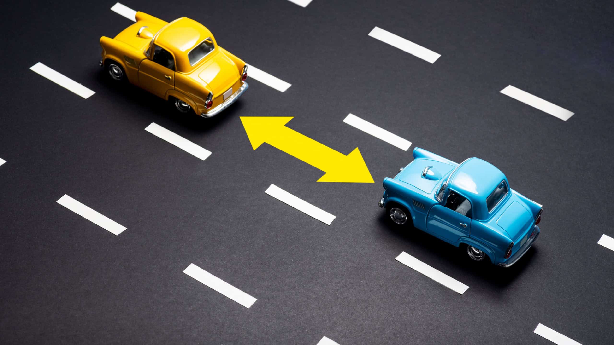 Two toy cars on the road one after another. One is blue one is yellow colored.