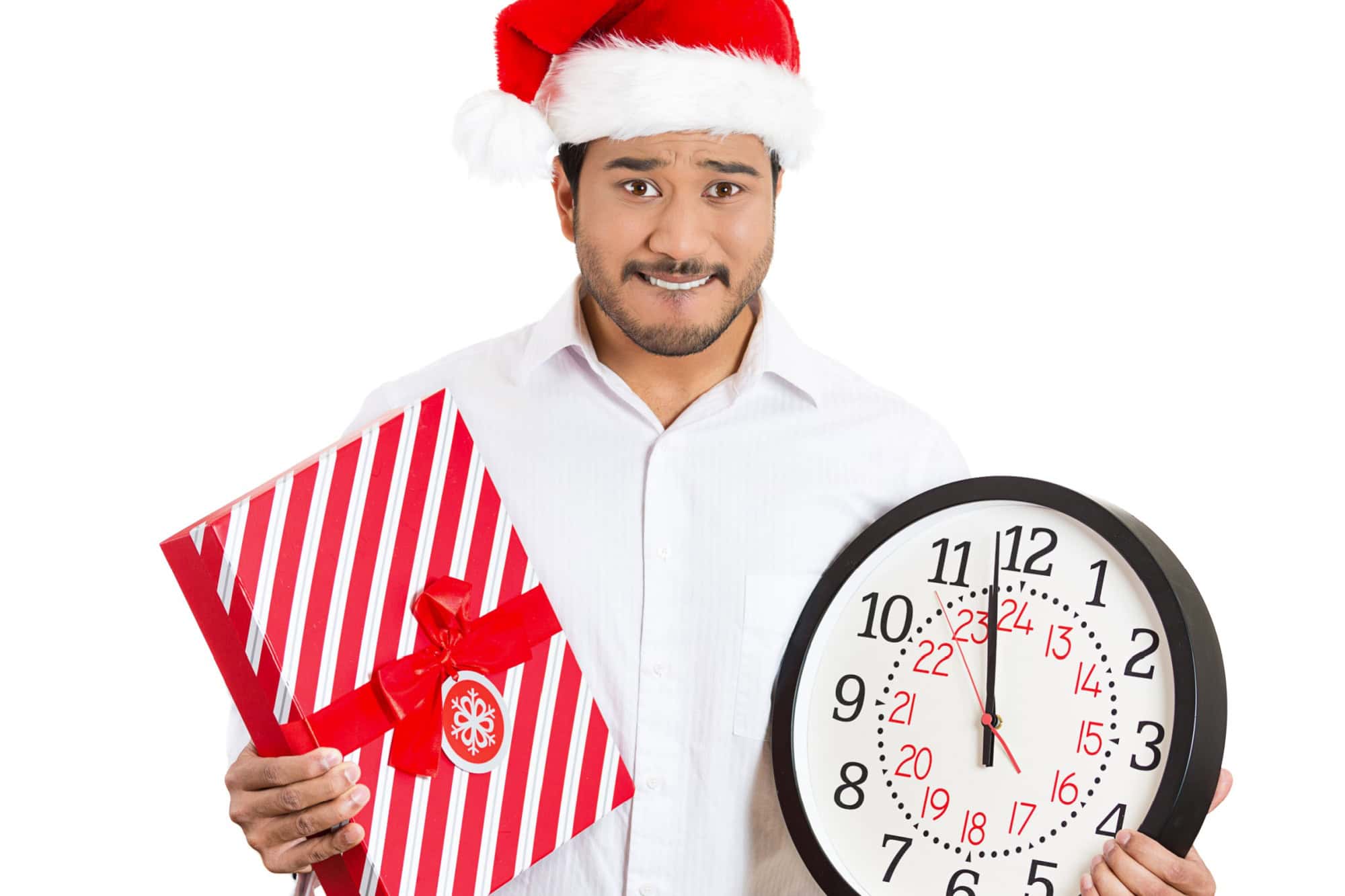 Closeup portrait of worried young man wearing red santa claus hat, holding clock and gift in hands, isolated on white background. Negative emotion facial expression. Last minute christmas shopping