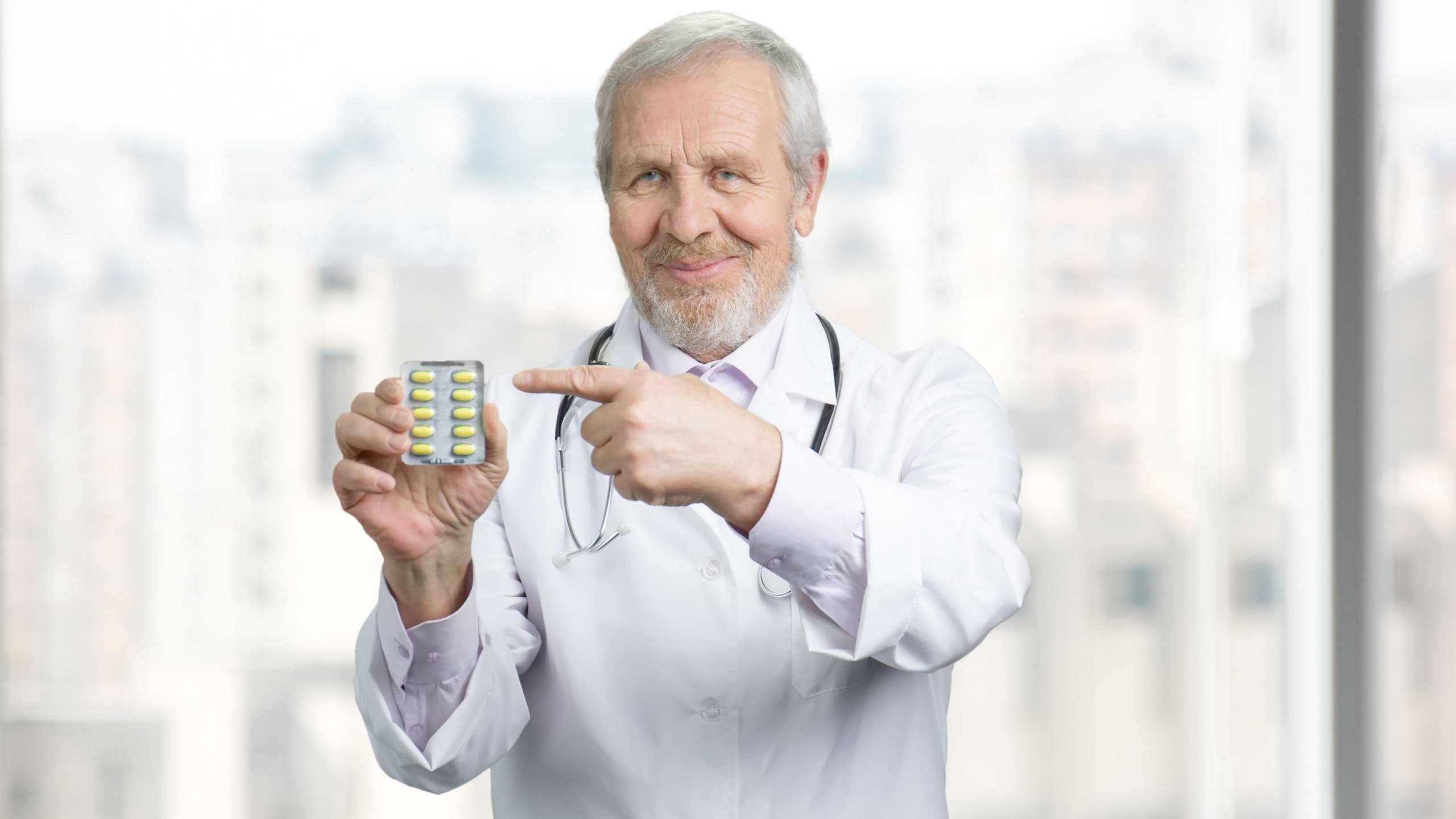 Old doctor pointing at pack of capsules. Concept of pills advertisement, recommending drugs medicine. Bright background.