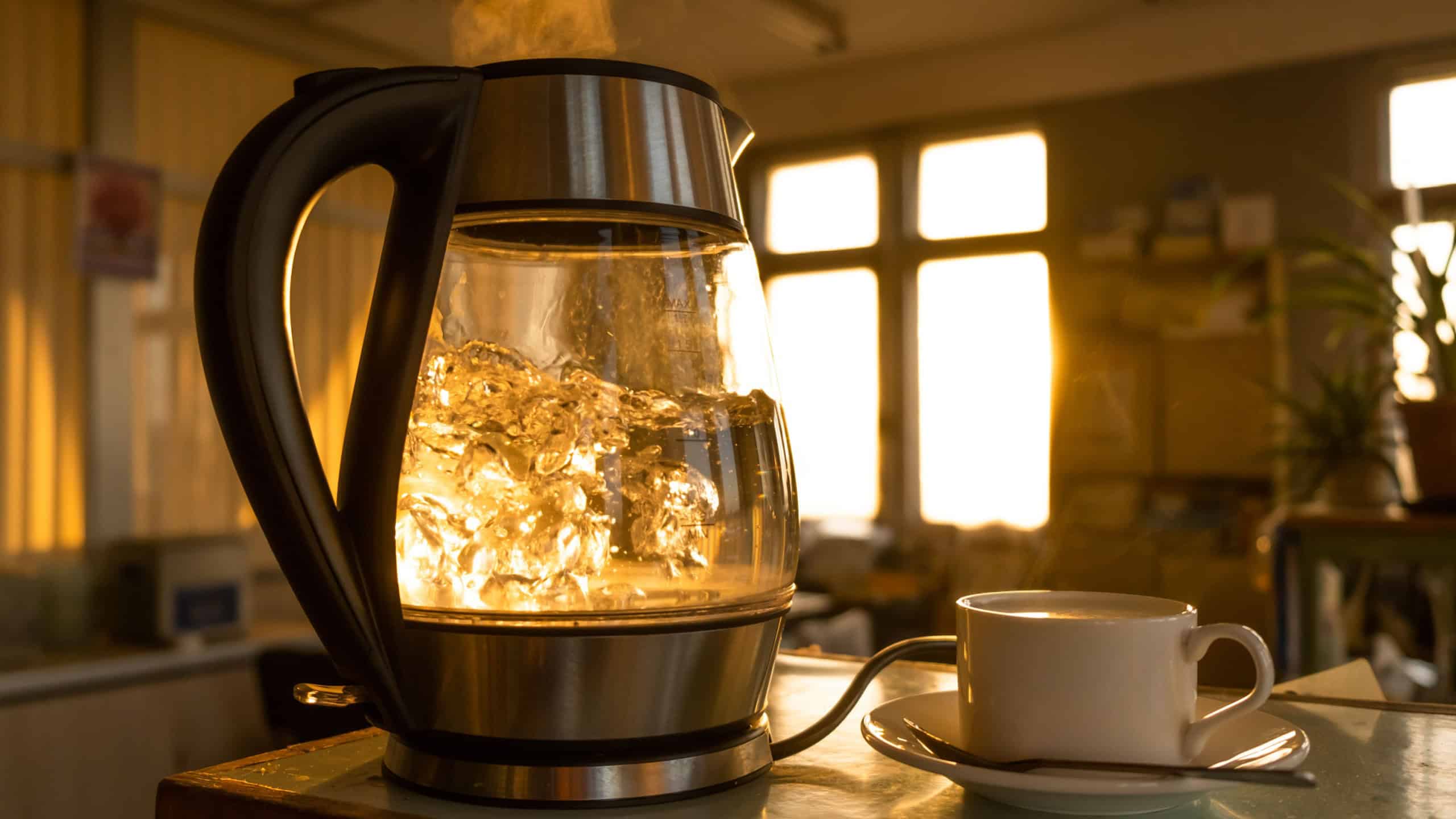 A transparent kettle of water boils against the background of the sunset shining through the window. The concept of coffee break and end of the working day.