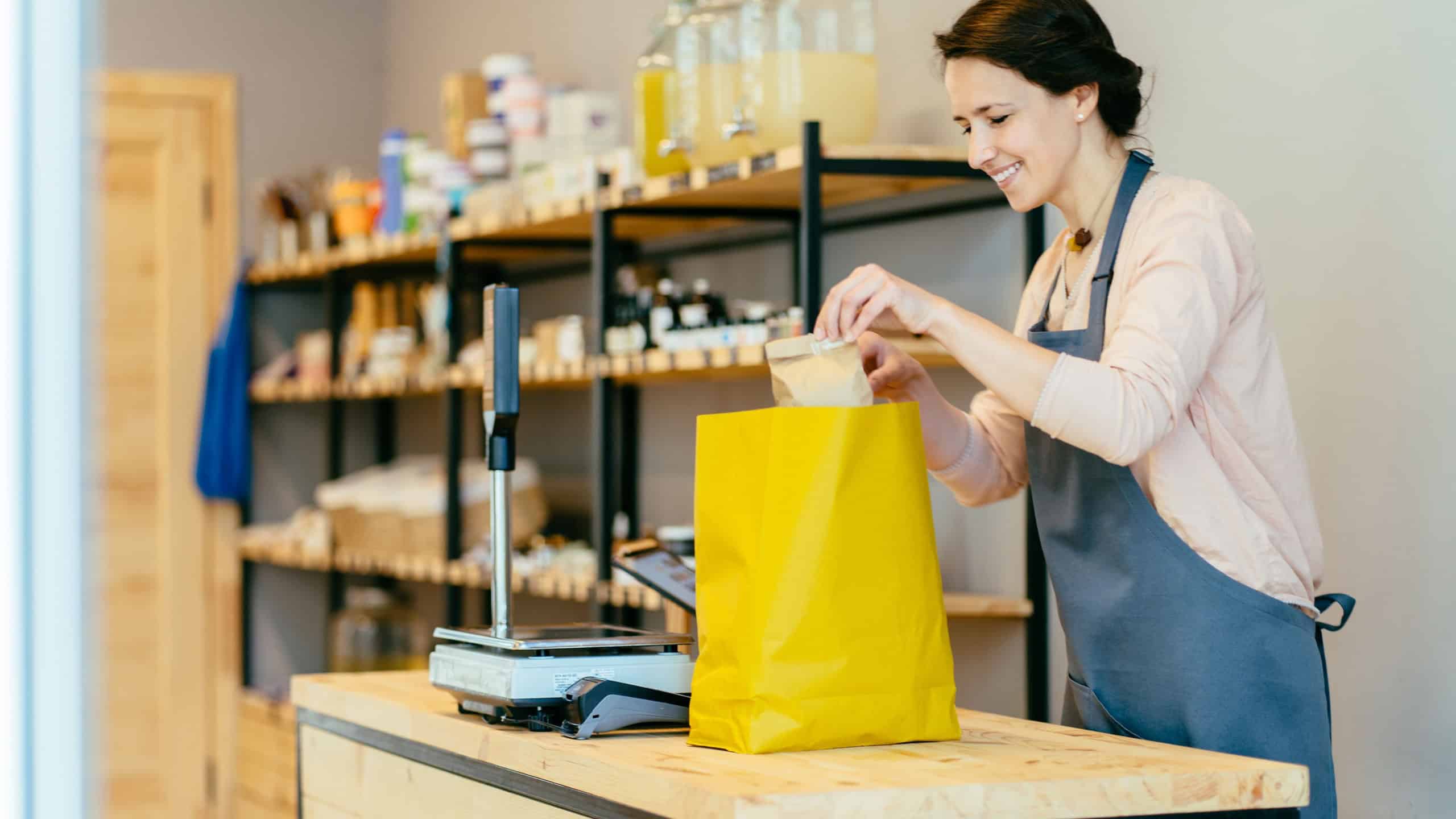Portrait of smiling shopkeeper in package free grocery store collects paper bag of food.Food delivery service. Female owner working behind counter at zero waste shop.Save planet, healthy life concept.