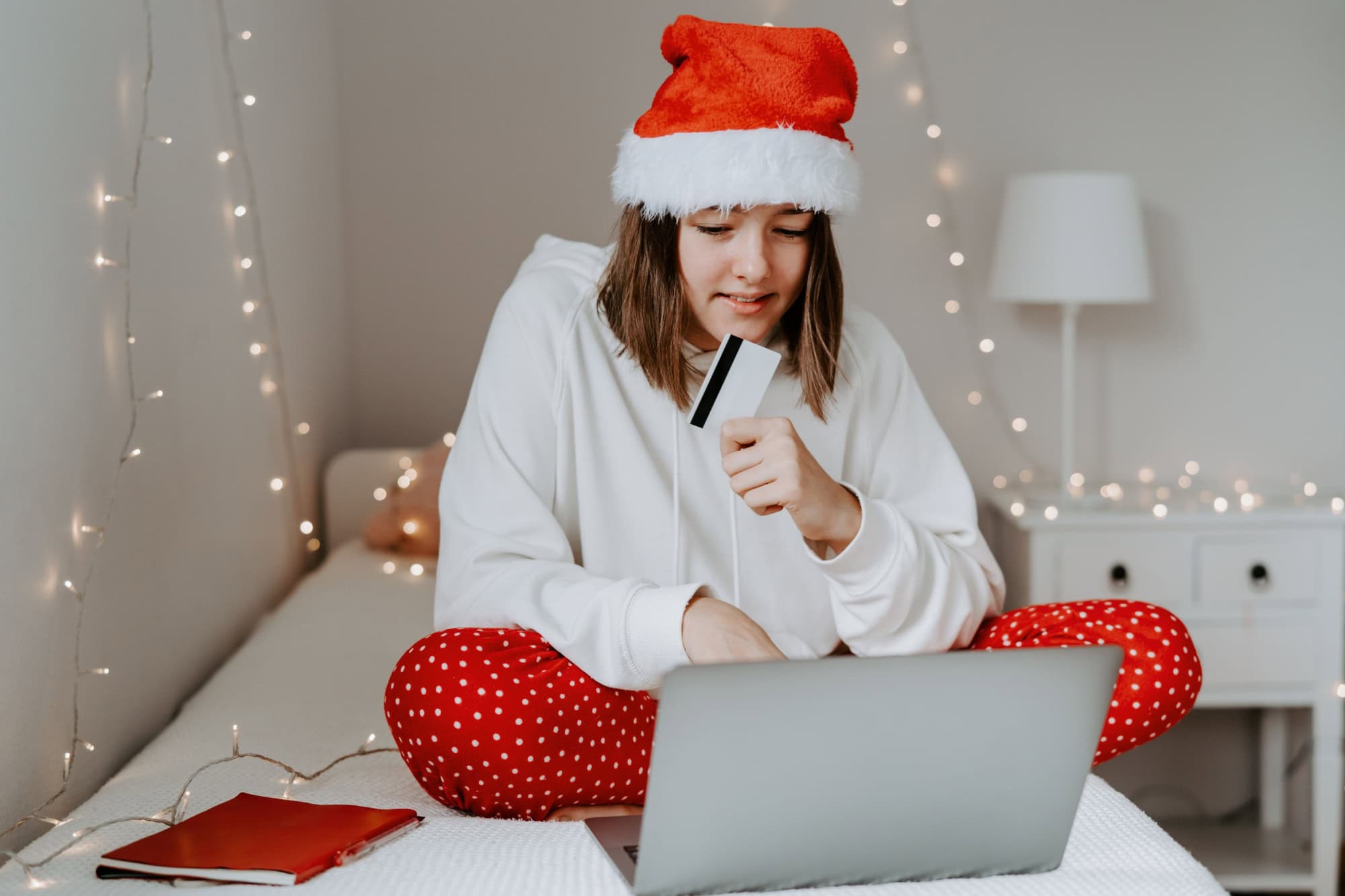 Young happy teen girl in red santa hat shopping online buying Christmas gifts paying by credit card using laptop sitting on bed in her bedroom