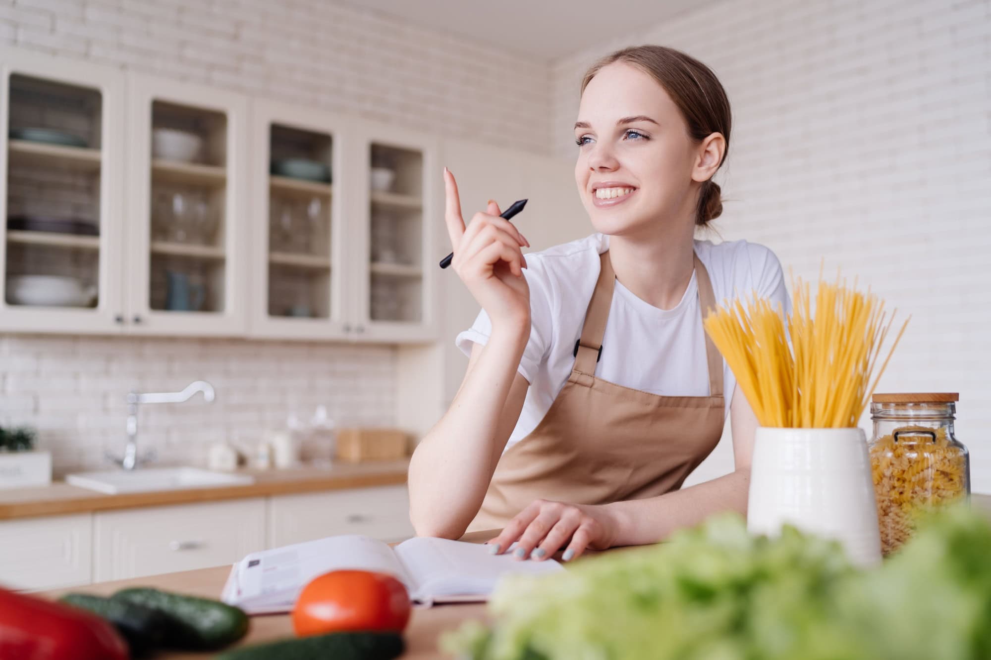 Young beautiful girl housewife in the kitchen in an apron, fresh vegetables on the table, writes down her favorite recipes, comes up with ideas for dishes