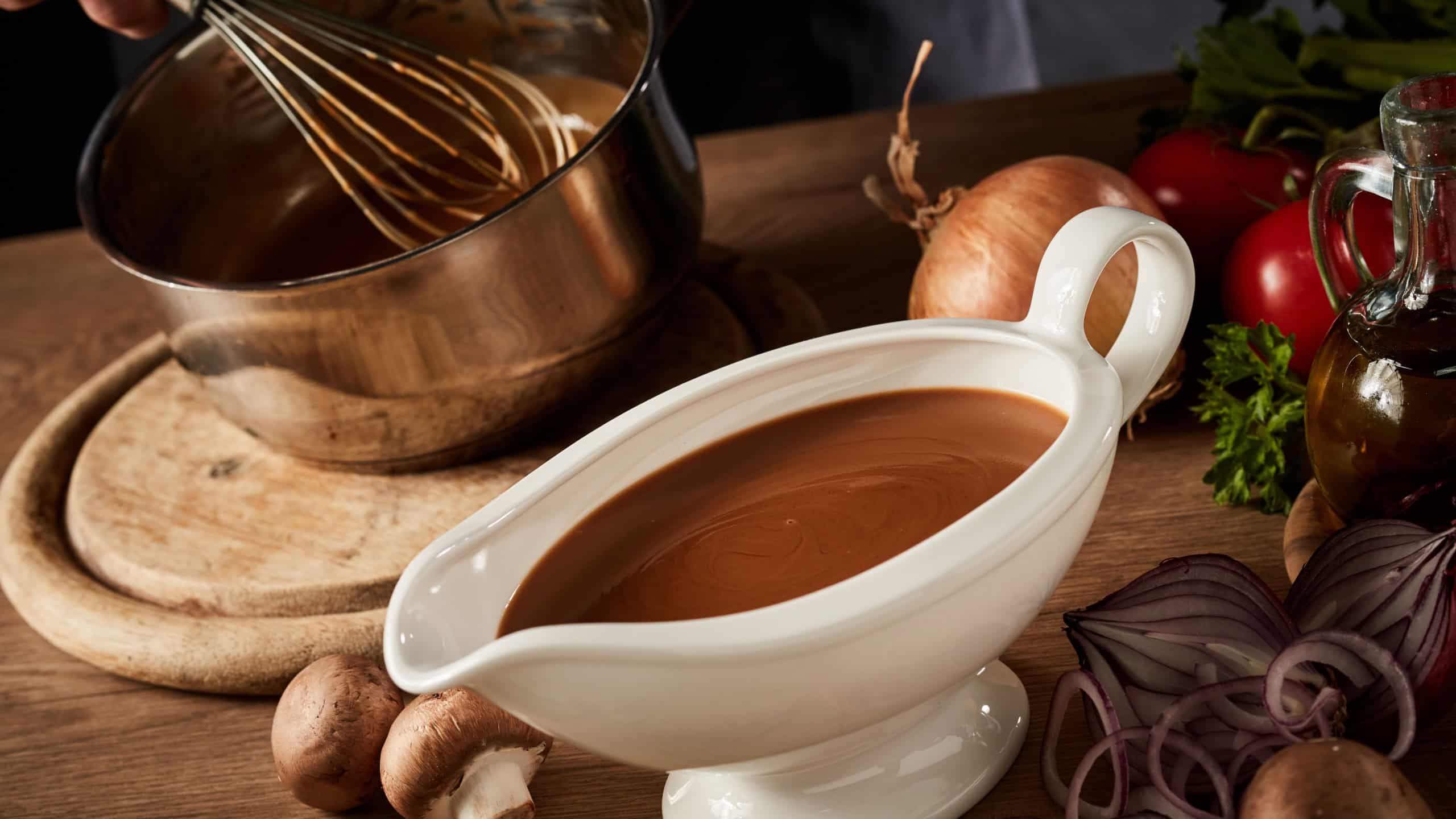 Sauce boat filled with rich brown spicy gravy viewed high angle on a kitchen table with assorted fresh vegetable ingredients