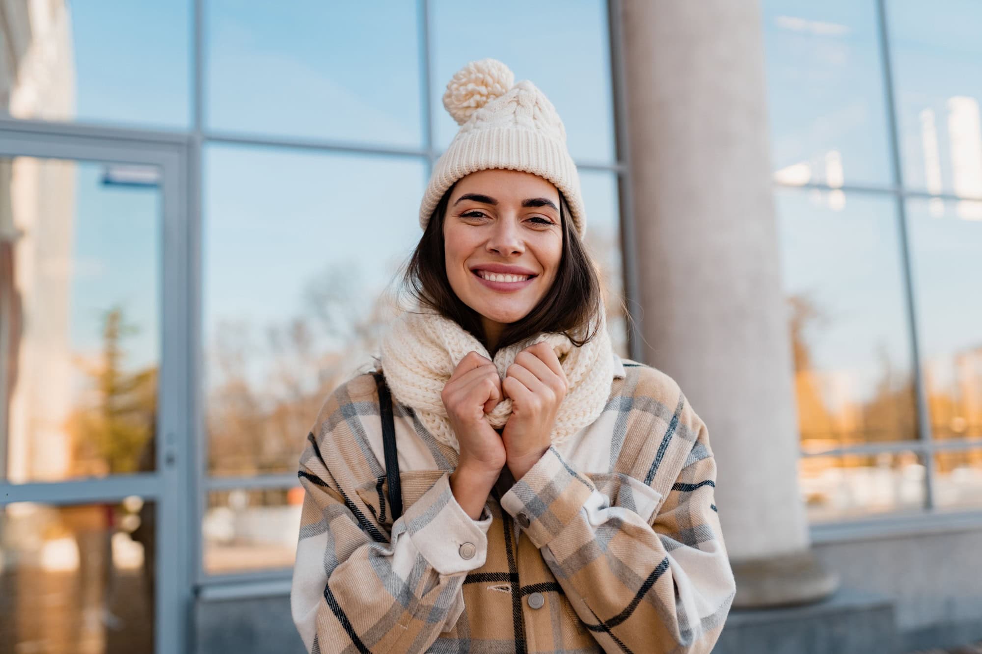 stylish attractive young smiling woman walking in street in winter outfit with coffee wearing checkered coat, white knitted hat and scarf, happy mood, fashion style trend