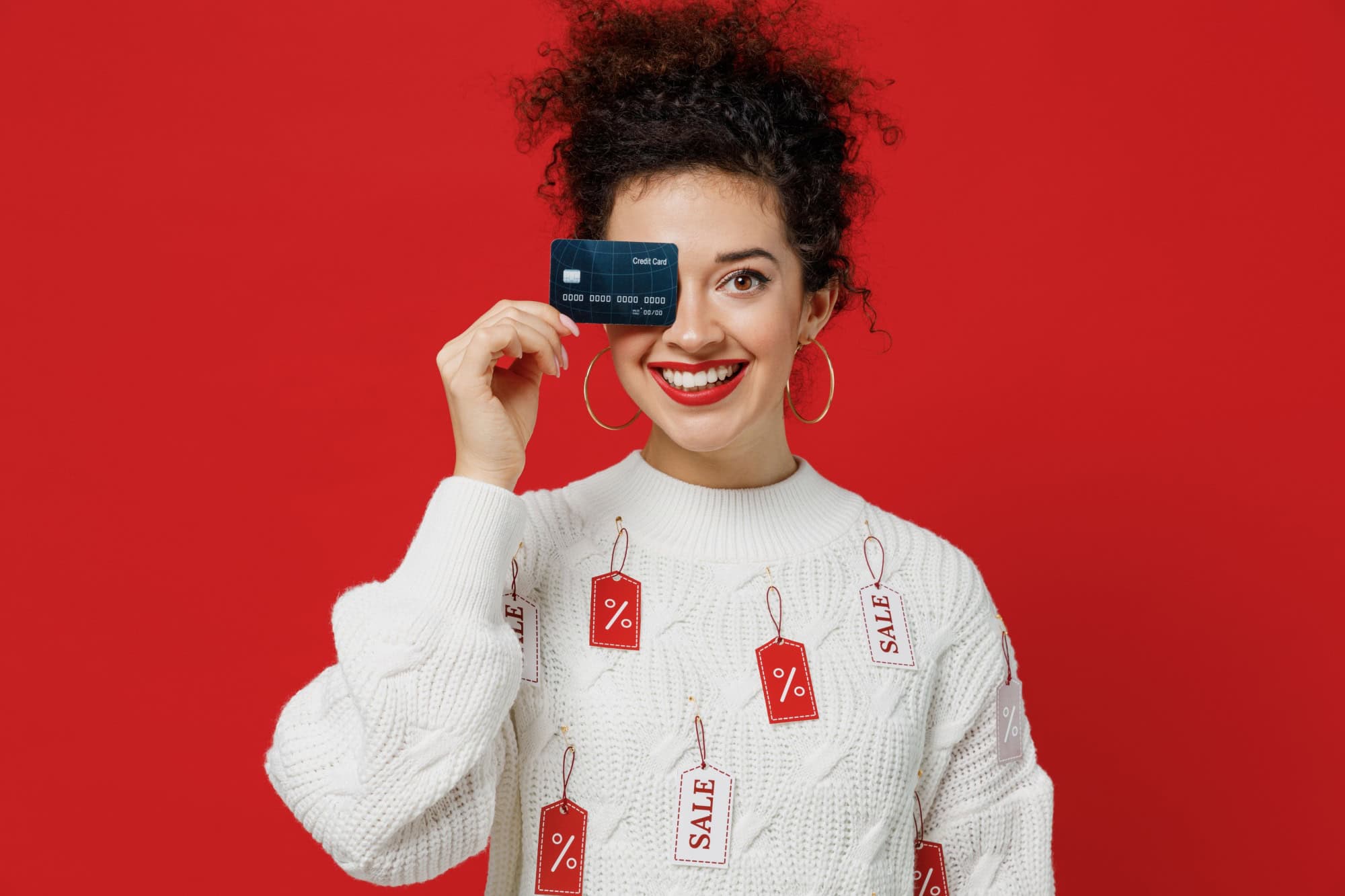 Young smiling happy rich female costumer woman 20s wear white knitted sweater with tags sale in store showroom hold cover eye with credit bank card isolated on plain red background studio portrait.