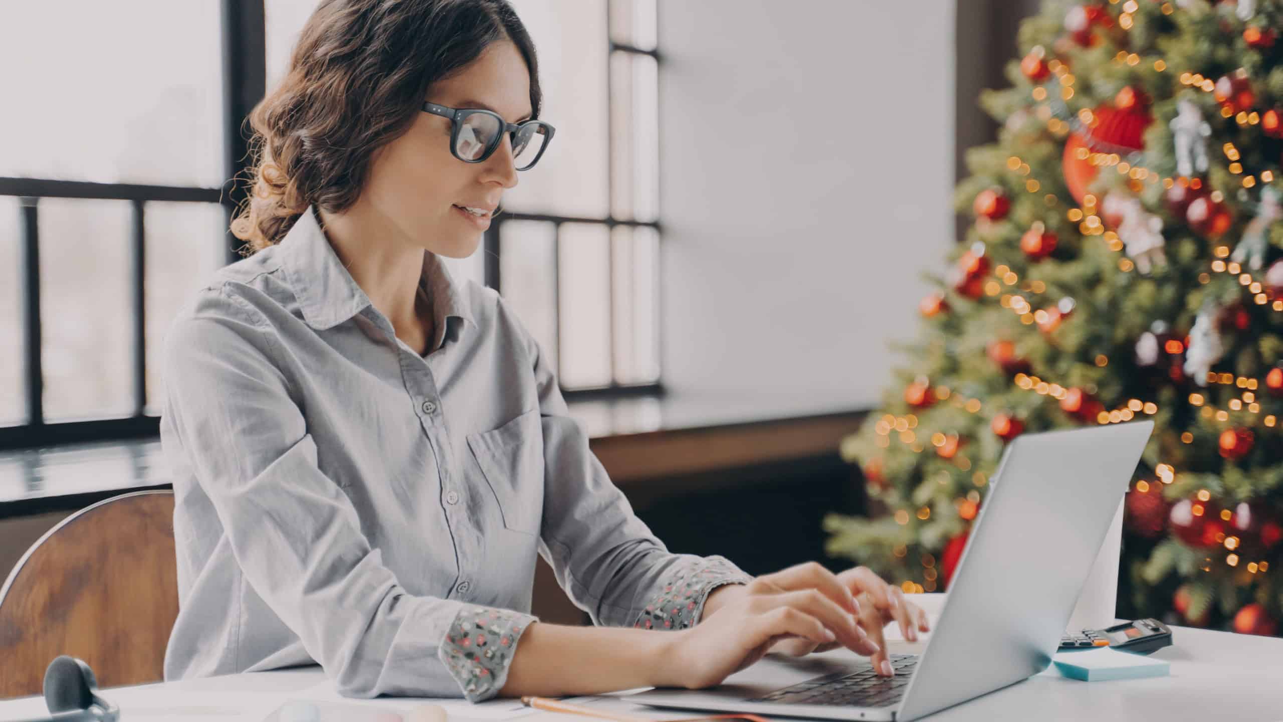 Businesswoman wearing glasses sitting in office near xmas tree and working on laptop during the holiday season, typing on computer keyboard. Hispanic female employee spending Christmas at work