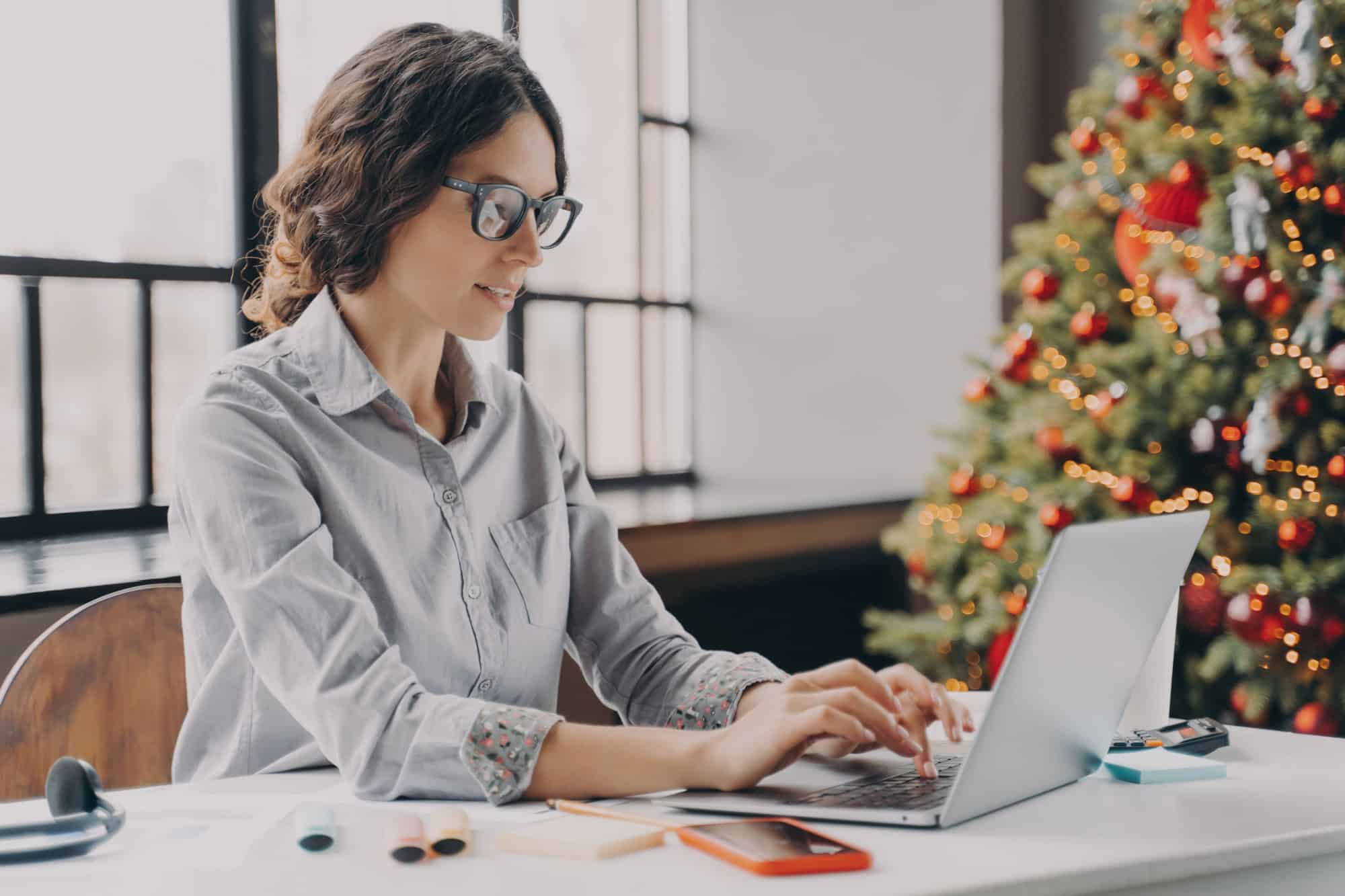Businesswoman wearing glasses sitting in office near xmas tree and working on laptop during the holiday season, typing on computer keyboard. Hispanic female employee spending Christmas at work