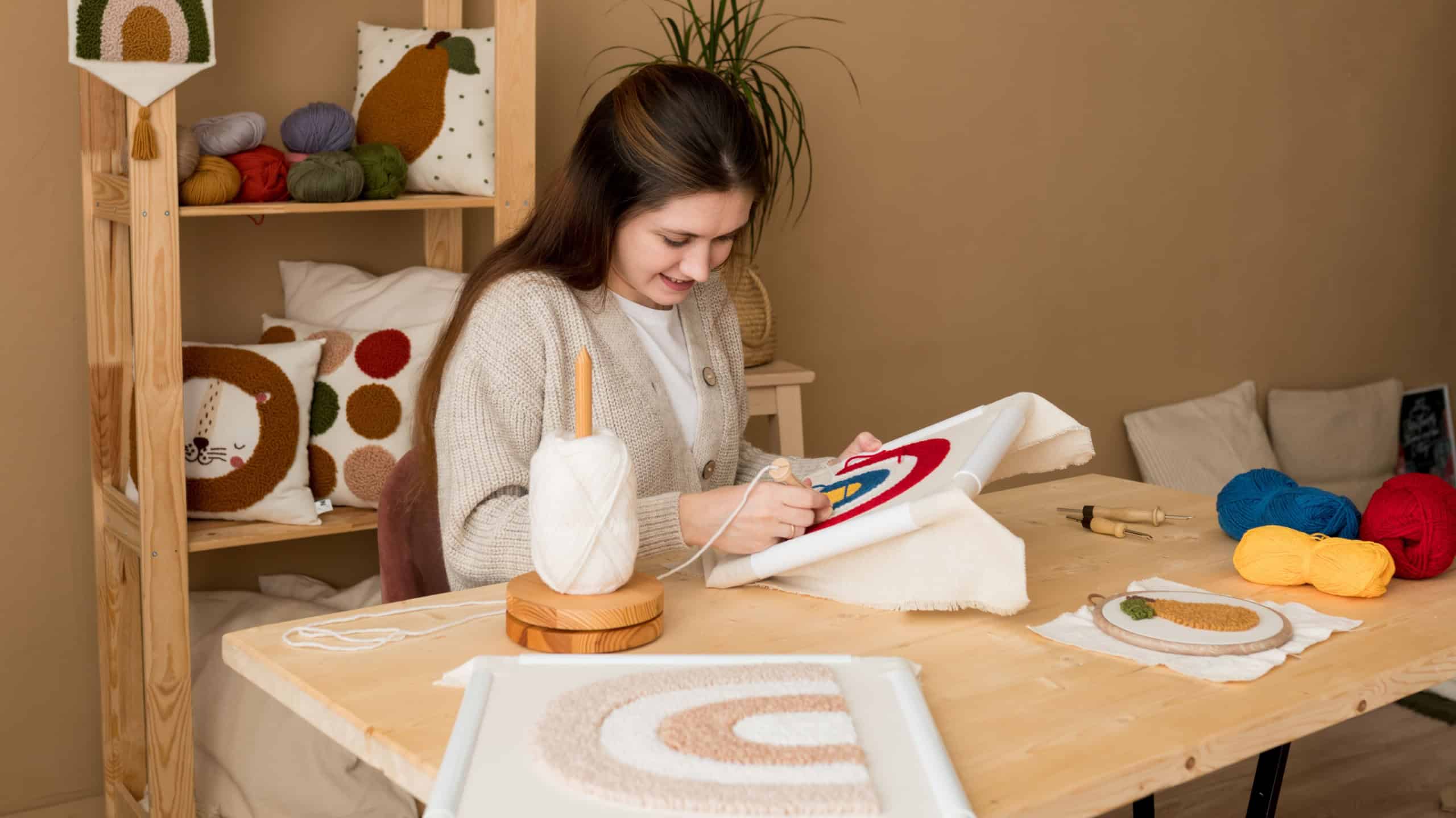 Punch needle embroidery. Young woman pushing the punchneedle straight down into the foundation fabric in workshop. Female embroidering handmade picture rainbow on canvas. Handicraft, hobby, diy