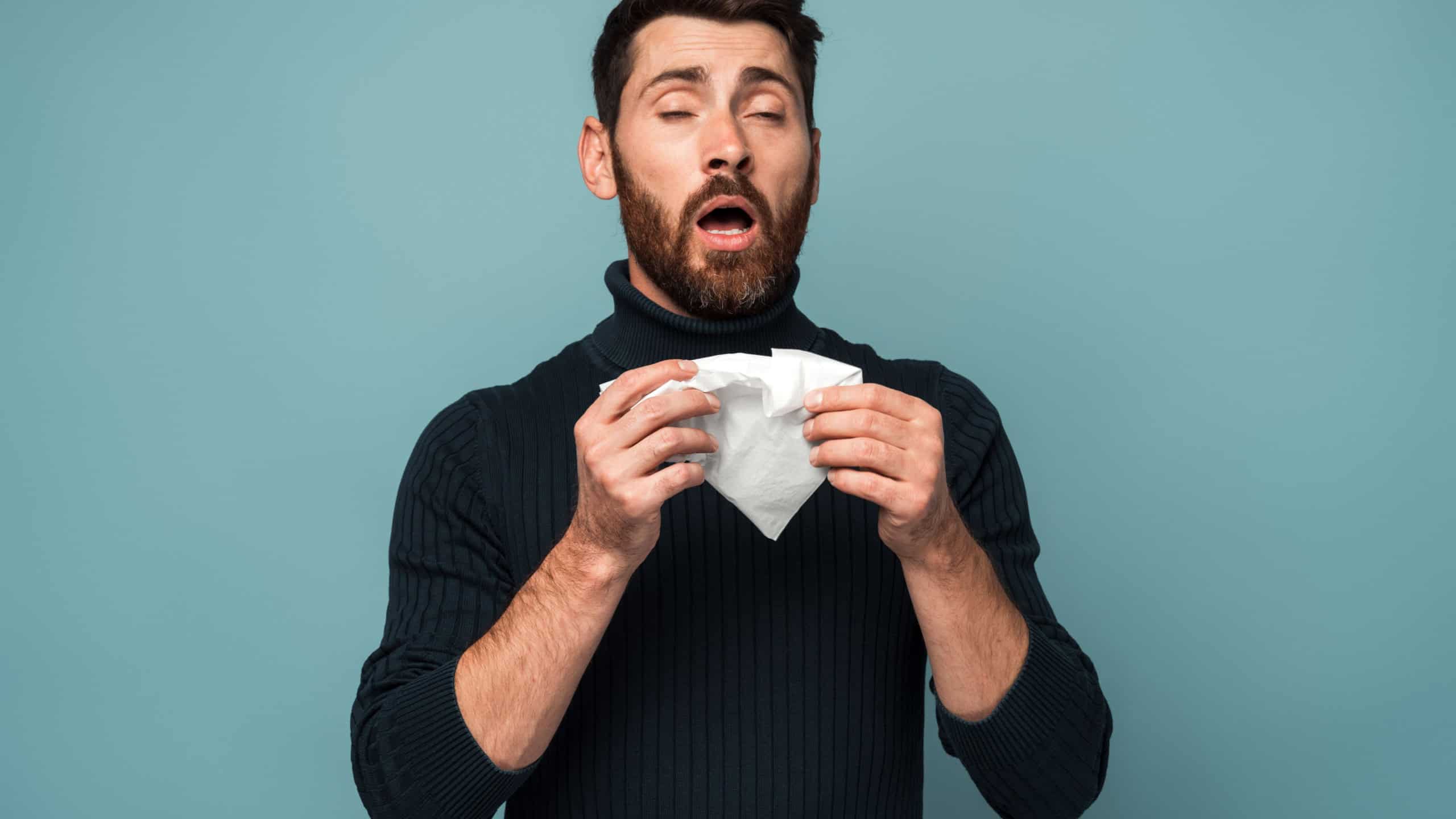 Influenza season. Unhealthy flu-sick man sneezing loudly in tissue, feeling unwell with runny nose, caught cold or allergy symptom. studio shot isolated on blue background 