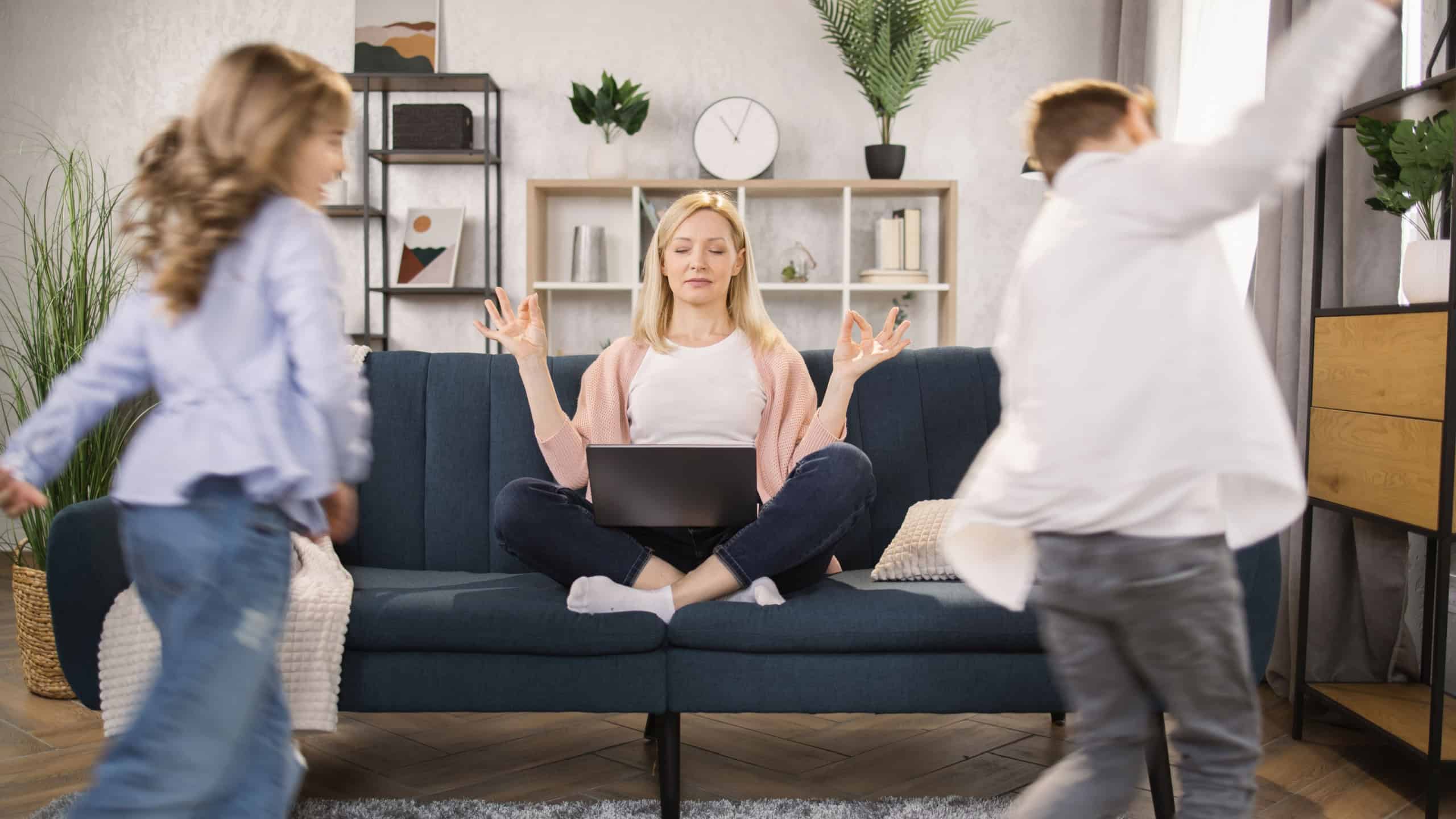 While noisy daughter and son kids run playing tag-game in living room near young caucasian mother she meditates sit in lotus pose on couch reached zen feel tranquil, stress relief self-control concept