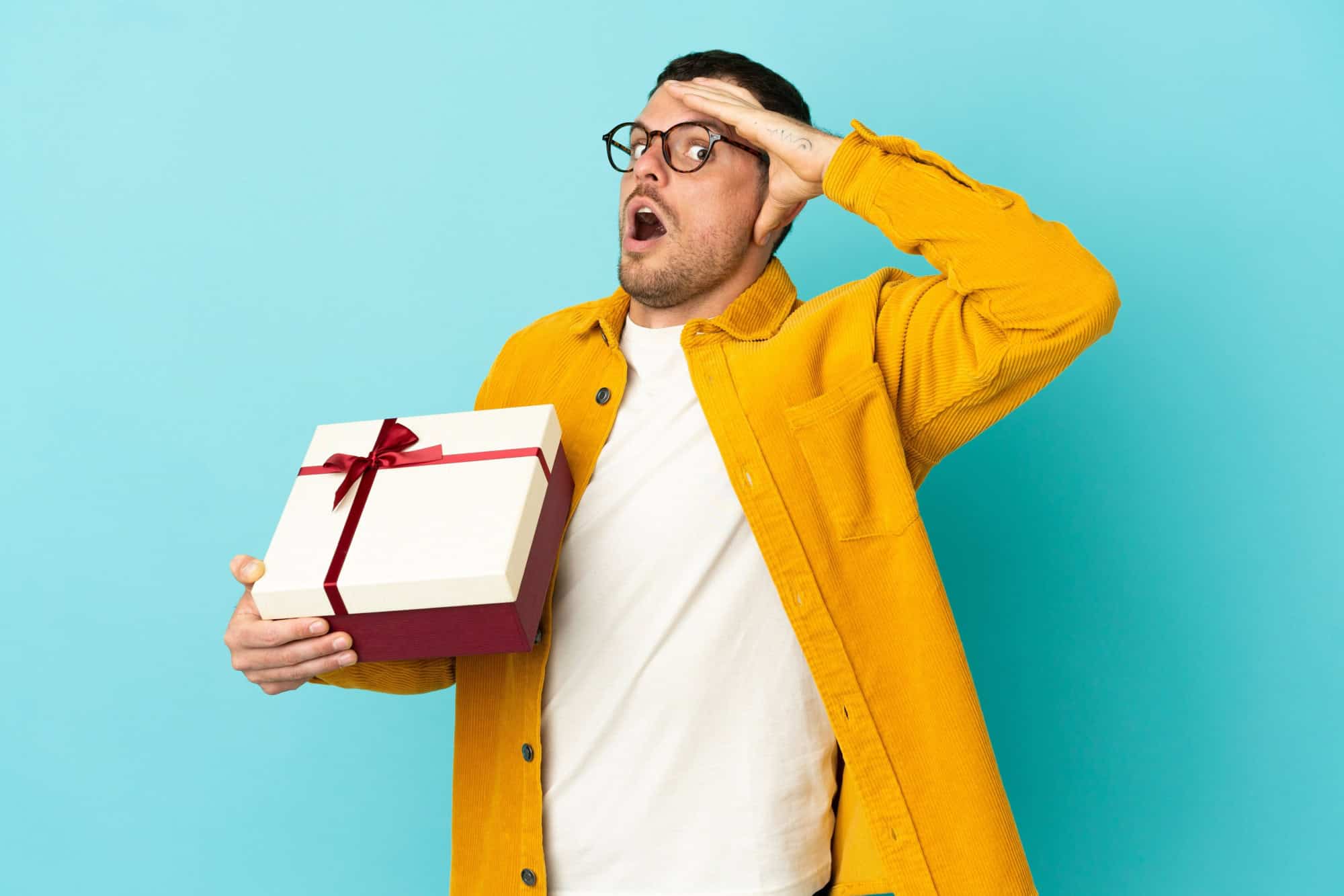 Brazilian man holding a gift over isolated blue background doing surprise gesture while looking to the side