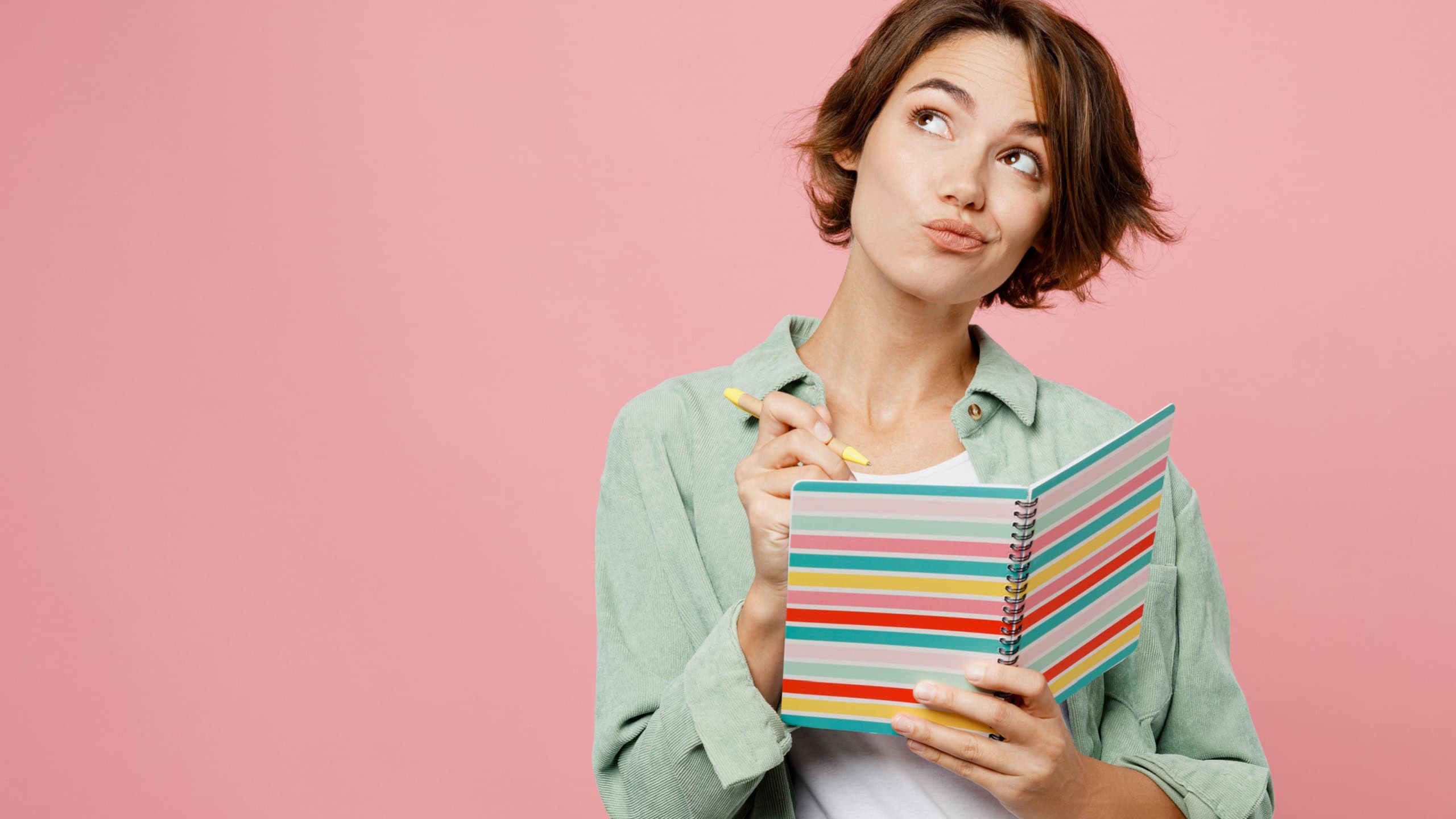 Young woman 20s she wear green shirt white t-shirt writing down in notebook diary remind memories and make list of dreams isolated on plain pastel light pink background studio People lifestyle concept