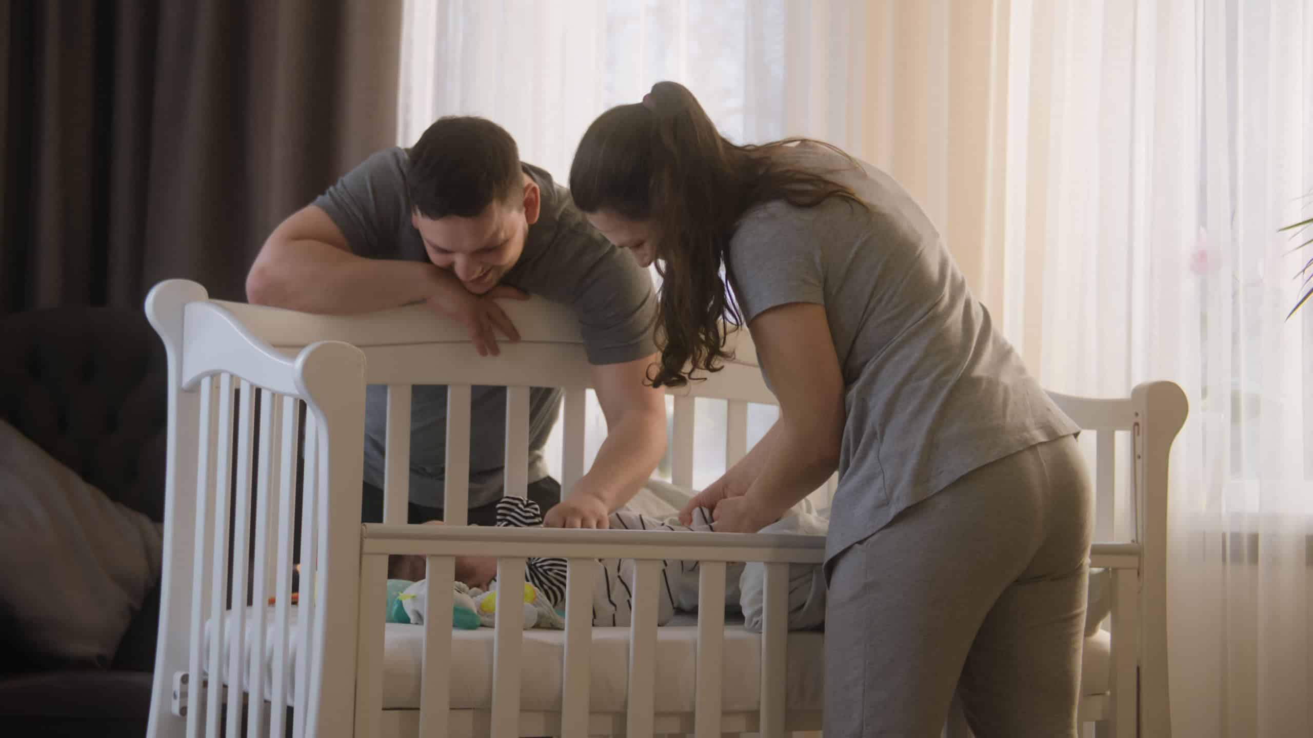 Father holds daughter or son in his arms and gives baby to wife. Loving mother puts baby in bodysuit in crib. Parents plays with newborn baby arms and legs. Concept of childhood, parenthood and family