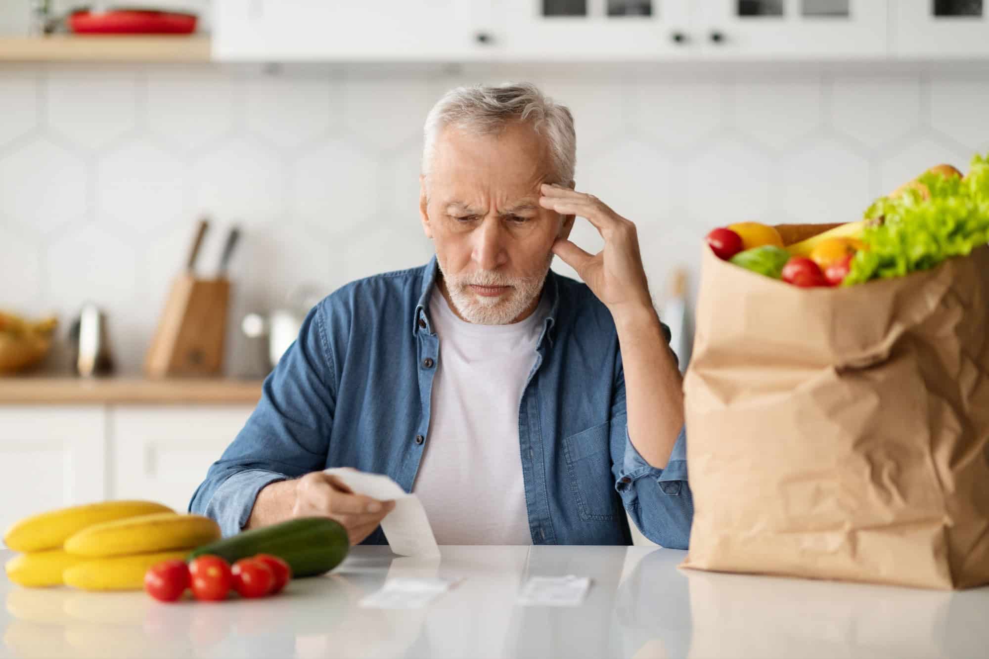 Grocery Expenses. Upset Senior Man In Kitchen Checking Bill After Food Shopping, Confused Elderly Gentleman Sitting At Table And Looking At Receipt, Suffering Financial Crisis And Poverty
