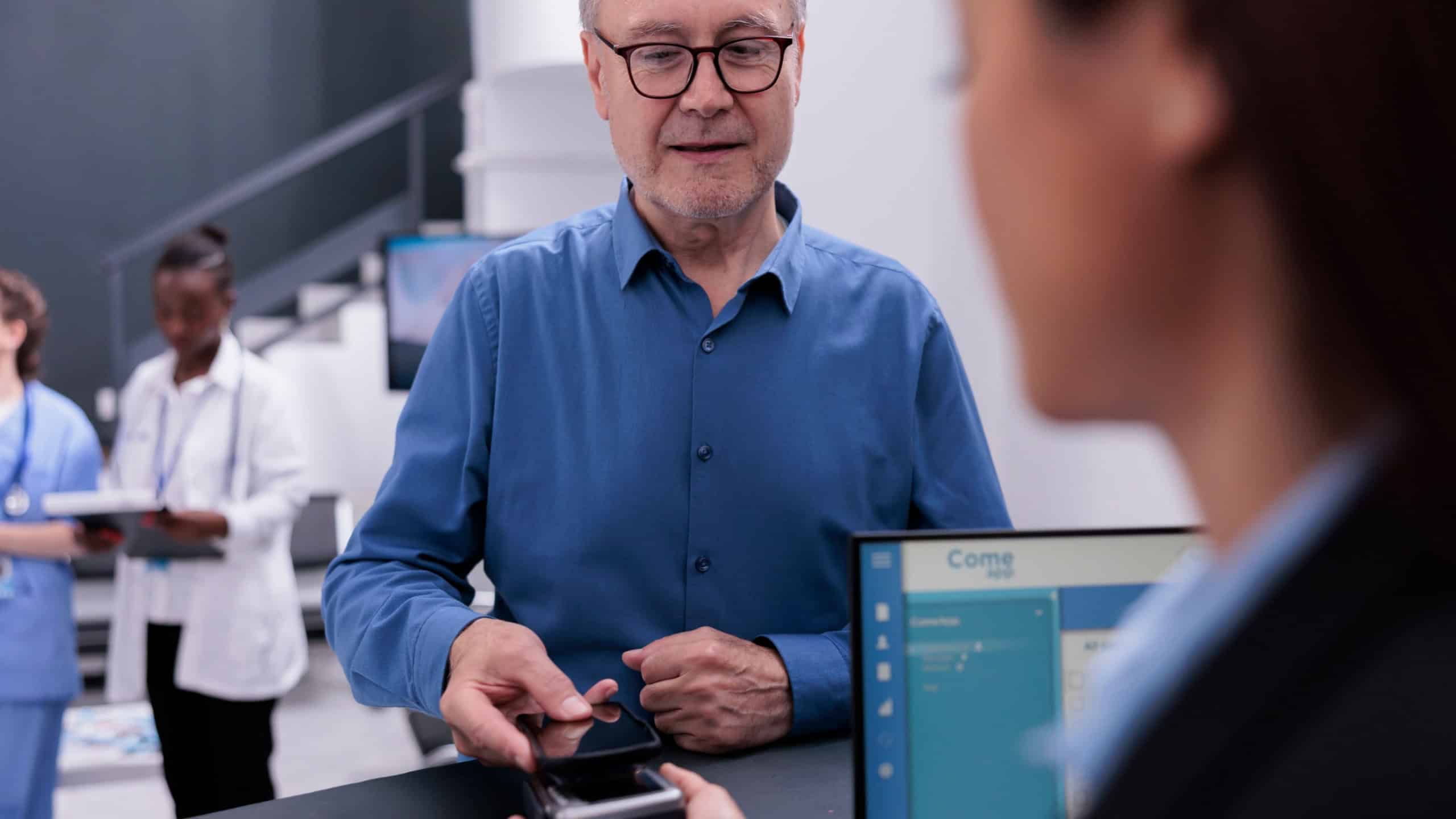 Elderly patient using phone contactless to pay checkup visit and prescription treatment at facility reception counter with receptionist. Man paying for examination appointment in waiting area lobby.