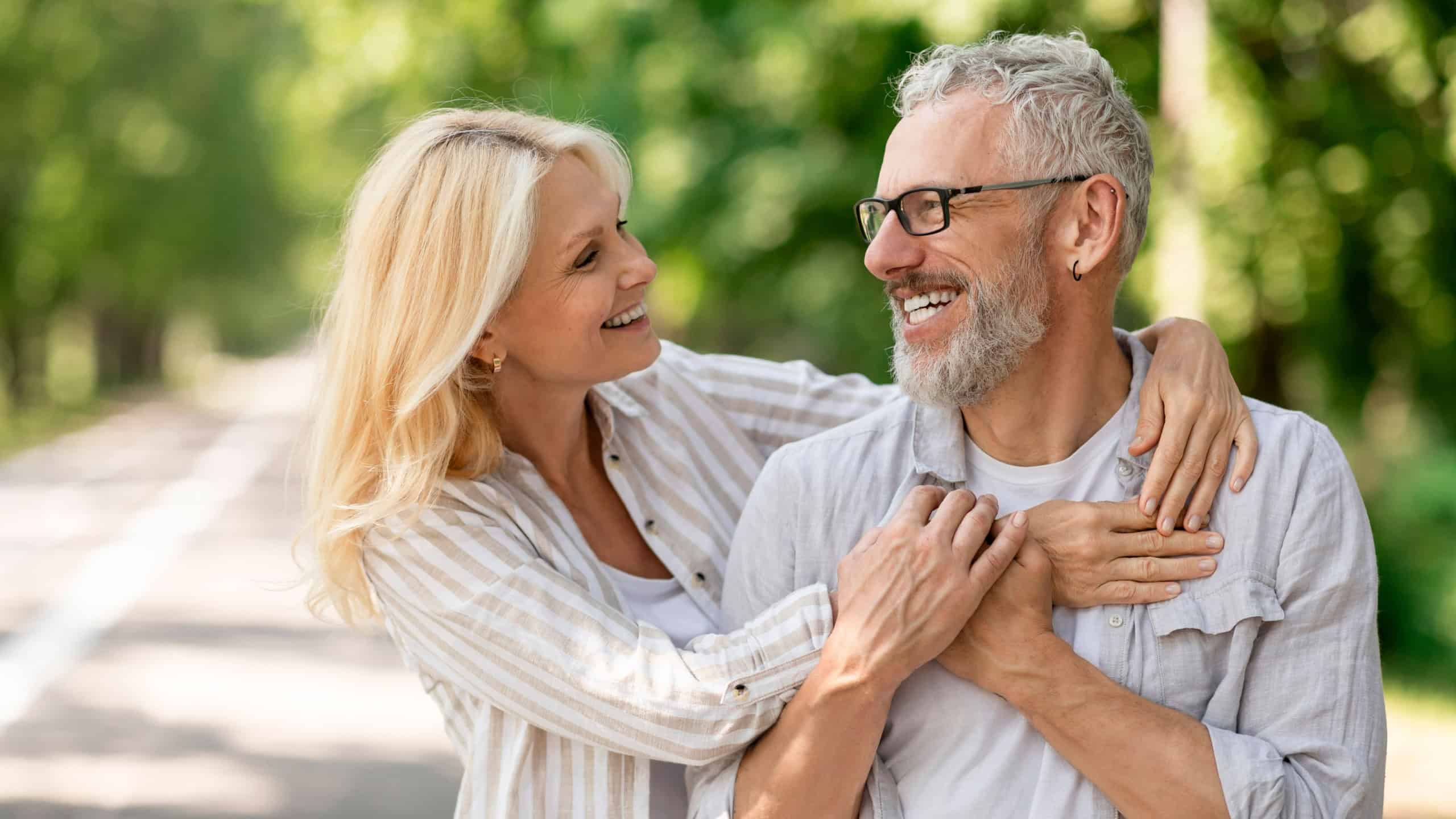 Happy romantic mature couple embracing while walking together in city park, smiling senior husband and wife hugging and looking at each other, cheerful elderly spouses enjoying date outdoors