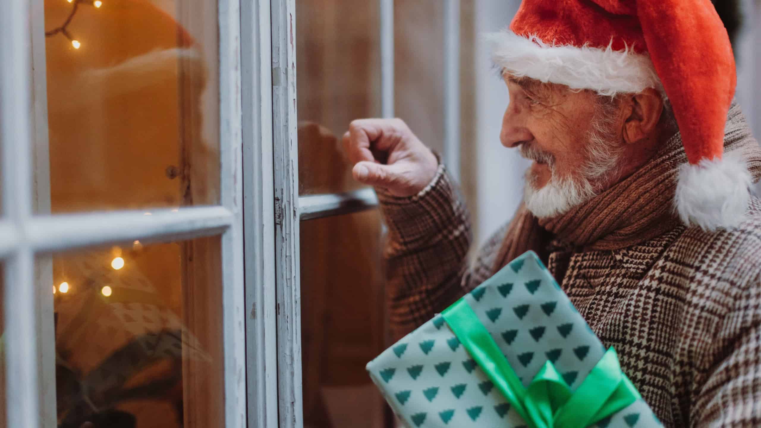 Senior man with a Christmas gift in hand standing in front of the window and knocking on it. Elderly man as Santa Claus with hat.
