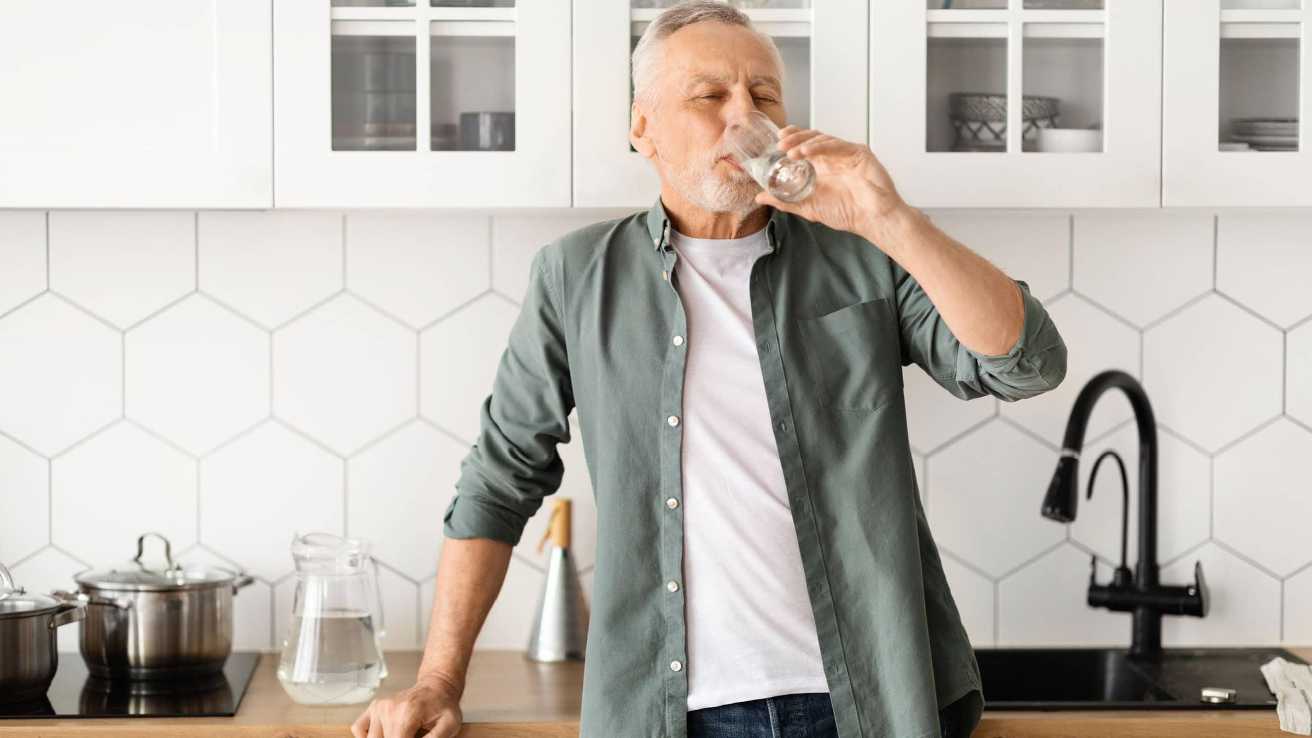 Healthy senior man drinking glass of water in his kitchen, elderly gentleman showcasing the importance of hydration for well-being, standing in relaxed posture, reflecting healthy lifestyle