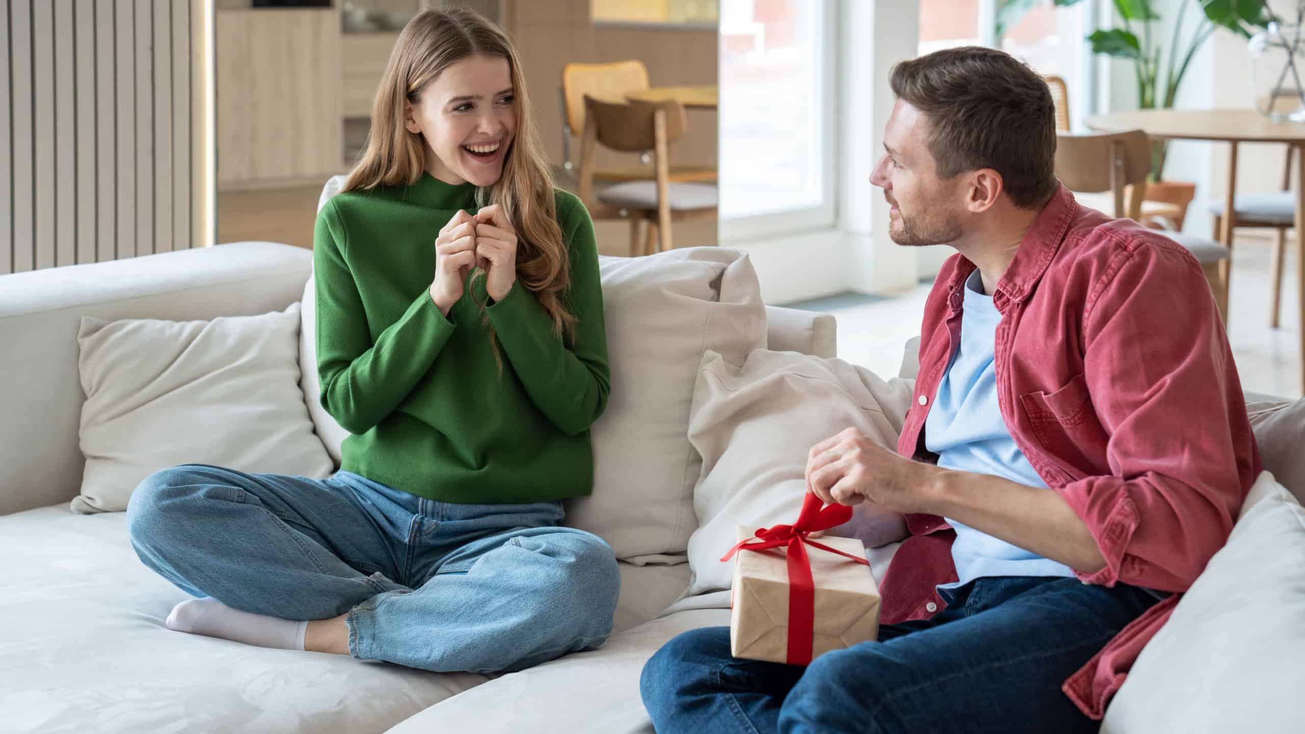 Joyful wife looks at husband unpacking present gift box smiling expecting reaction sit on couch at home. Woman congratulating man. Happy couple celebrating birthday anniversary family holiday together