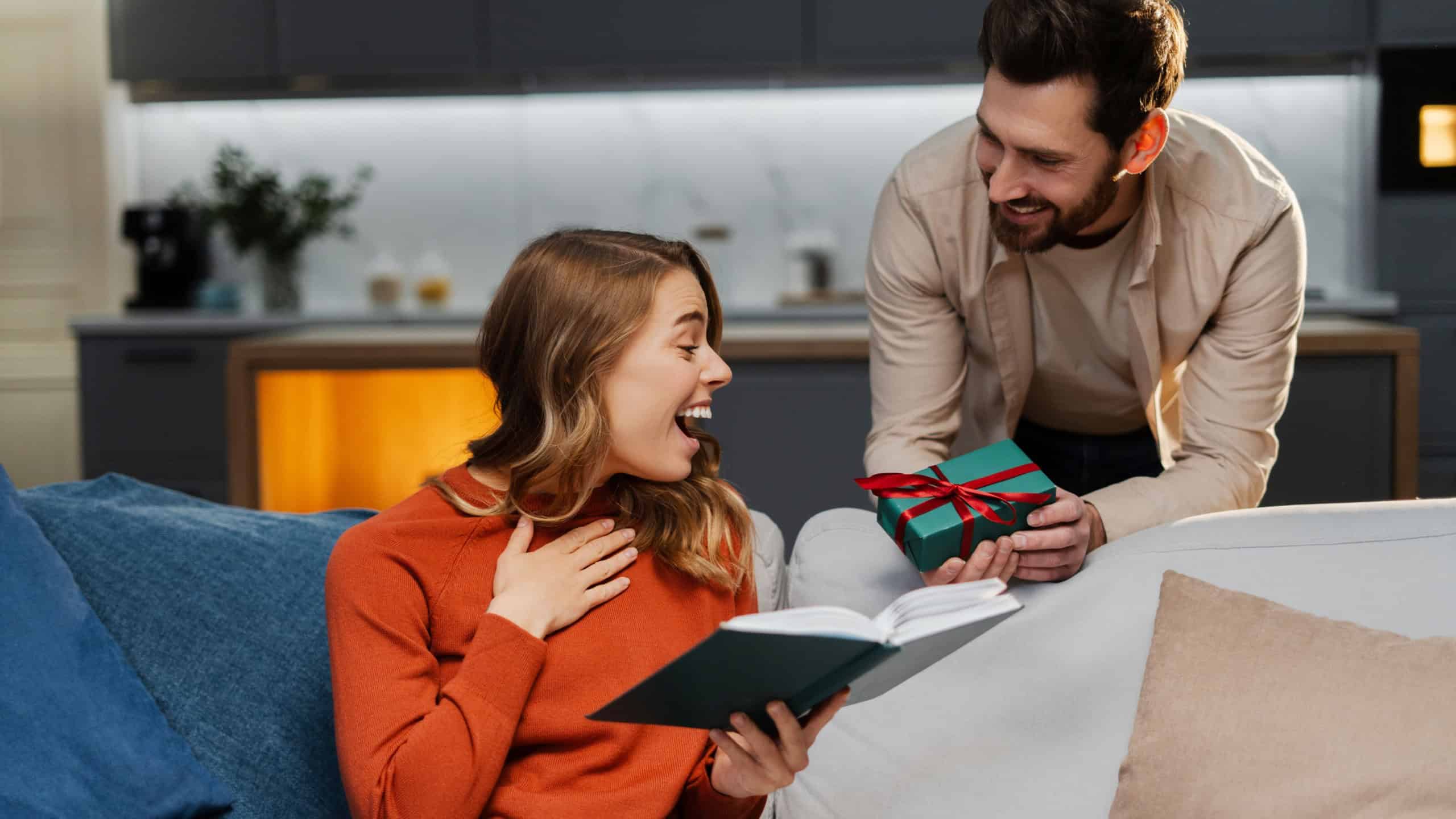 Young happy couple holding gift box celebration holiday at home. Excited beautiful woman looking at present sitting on sofa. Birthday concept