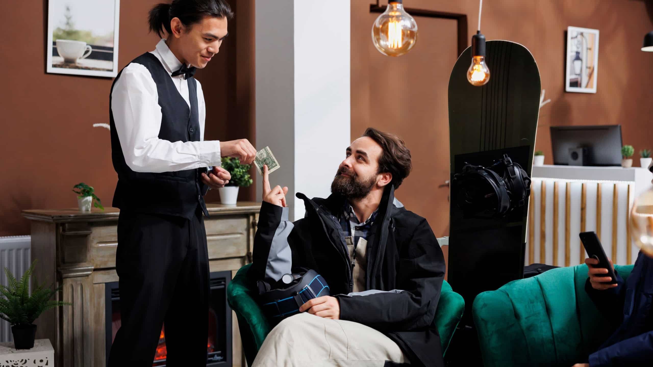 In hotel reception, friendly waiter caters to couple with skiing and snowboarding equipment. While woman is browsing on her cell phone, young man gives money tip to employee in luxury winter resort.