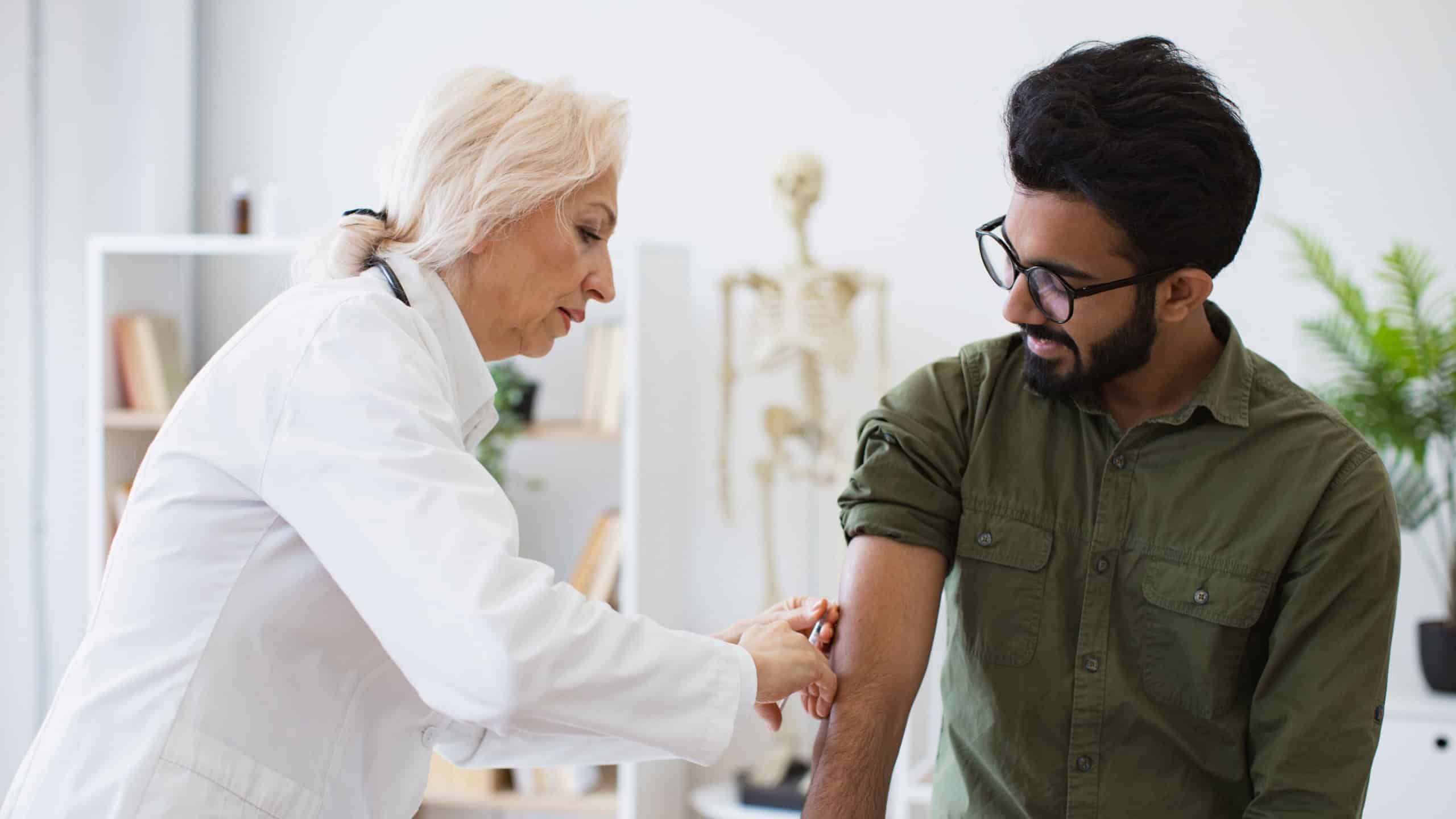 Confident mature woman in lab coat holding syringe with seasonal flu vaccine while young man sitting in exam room. General practitioner giving injection to boost immunity for young bearded patient.