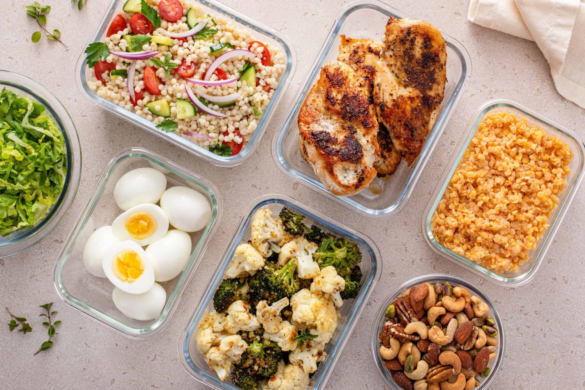 Healthy meal prep with cooked chicken breast, boiled eggs, roasted vegetables, cooked lentils, couscous salad and mixed nuts
