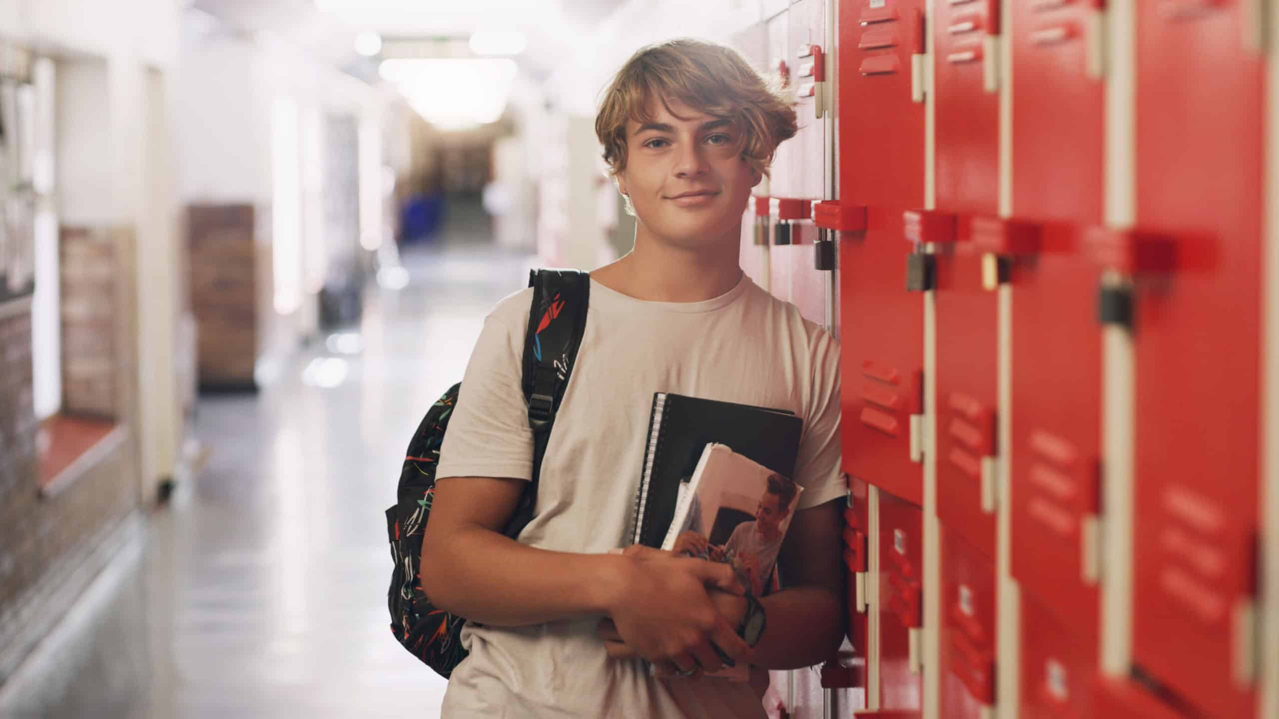 Boy, portrait and hallway as high school student by lockers with books for education, scholarship or campus. Male person, face and learning development with backpack in corridor, academic or study