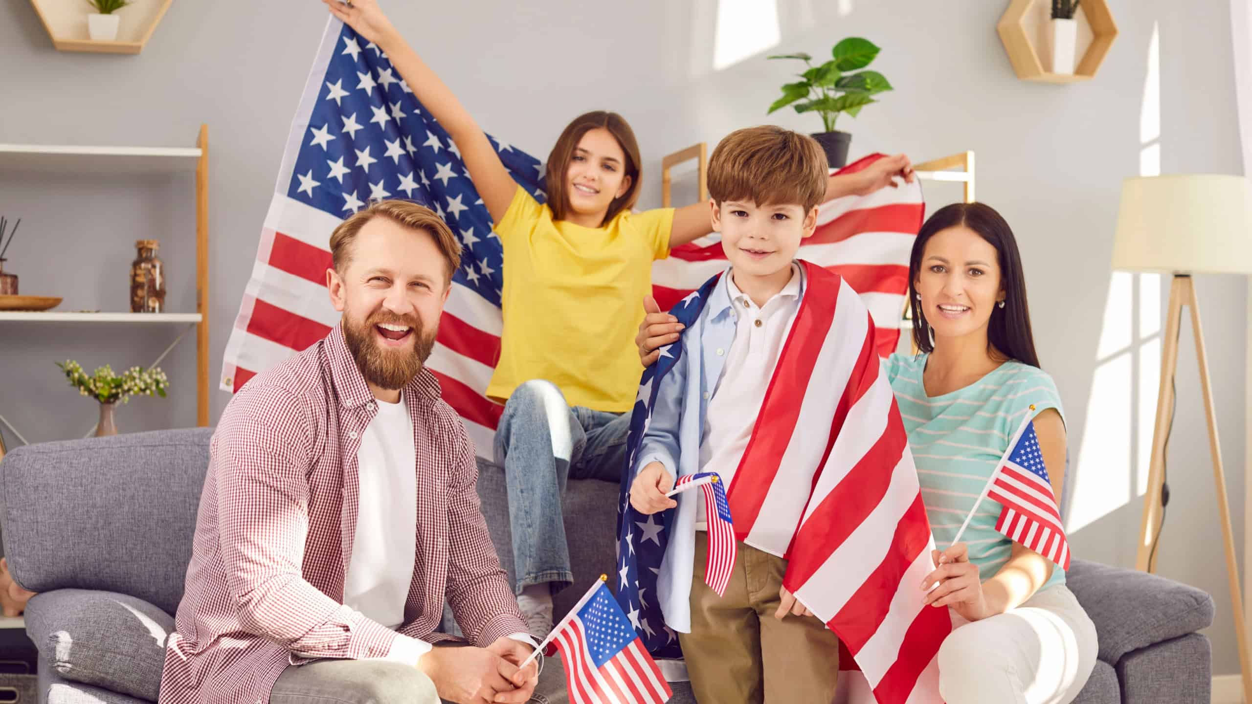 Portrait of a young happy American family with two kids sitting on sofa at home with flag of united states and looking cheerful camera celebrating Independence Day. Patriotic US holiday.