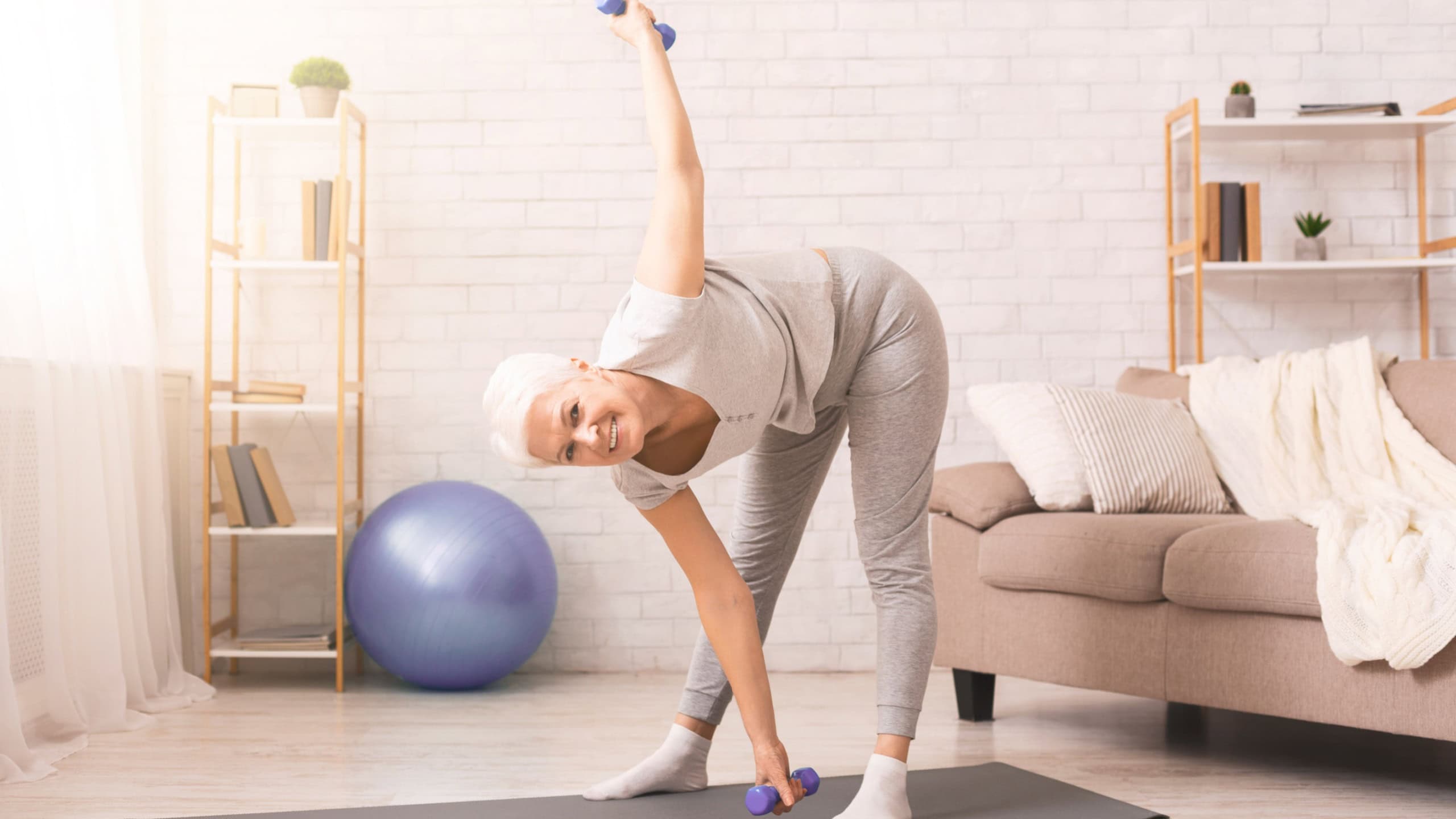 A smiling elderly woman is engaging in a fitness routine, holding a set of blue dumbbells while performing a side bend. She is standing on a yoga mat in a well-lit living room
