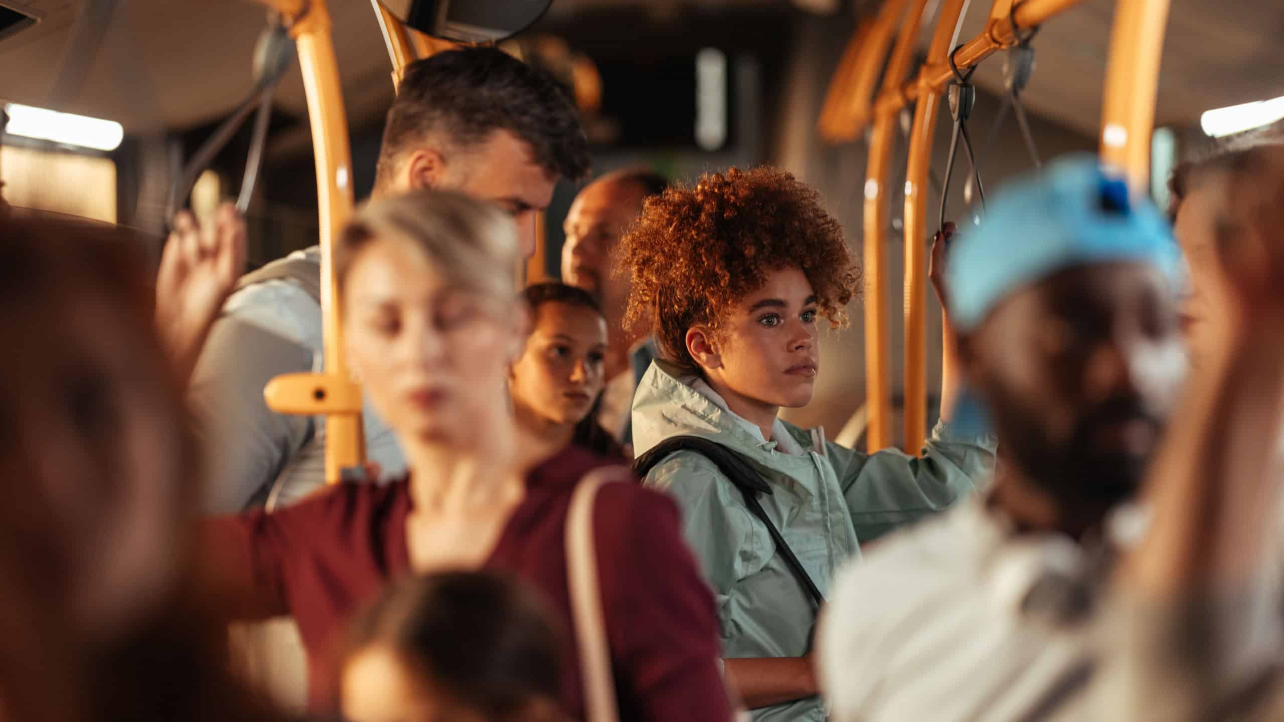 Teenage girl with curly hair standing on a crowded public bus among other passengers.