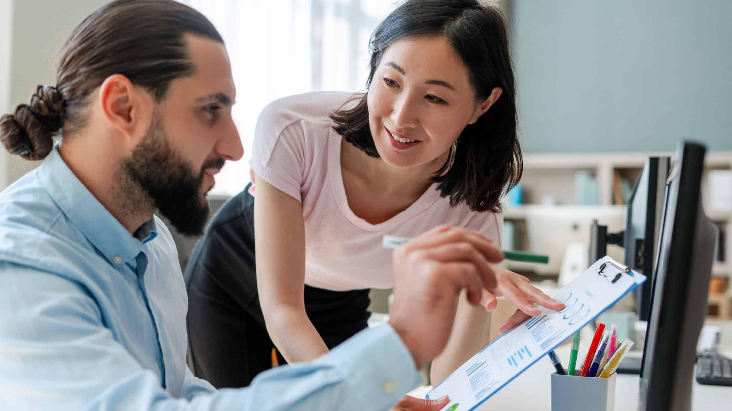 Smiling successful colleagues, businessman and businesswoman, Arab man and Asian woman working together in modern office, near desk. Teamwork concept