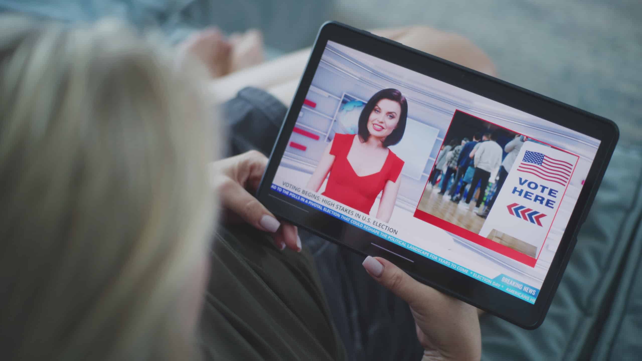Close up of TV news broadcast with female news anchor displayed on tablet screen. Caucasian woman watches breaking news about US presidential elections using tablet computer while lying on sofa.