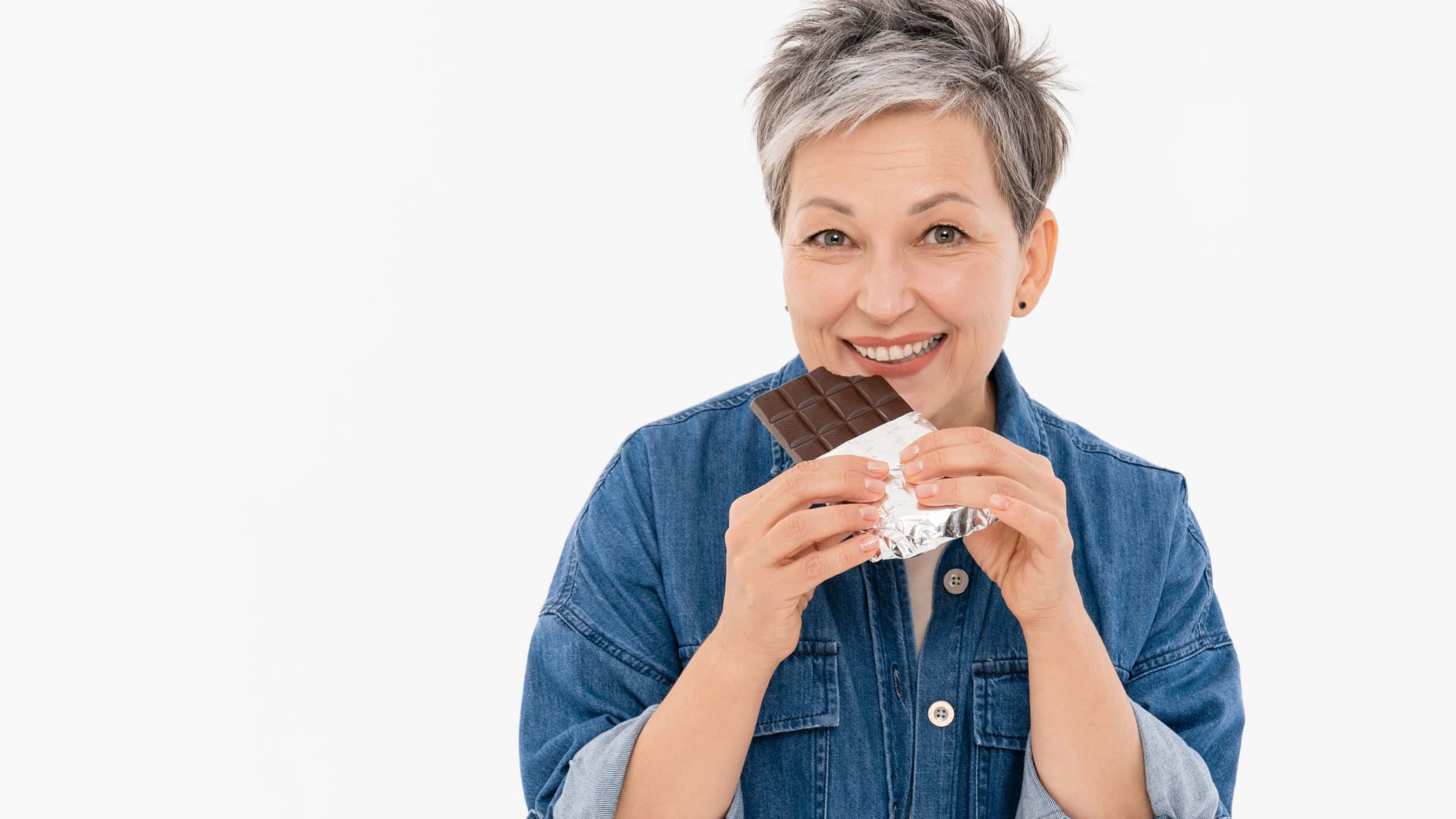 Close up portrait of middle-age woman lady with short grey hair in casual outfit white t-shirt denim eating tasty yummy chocolate bar