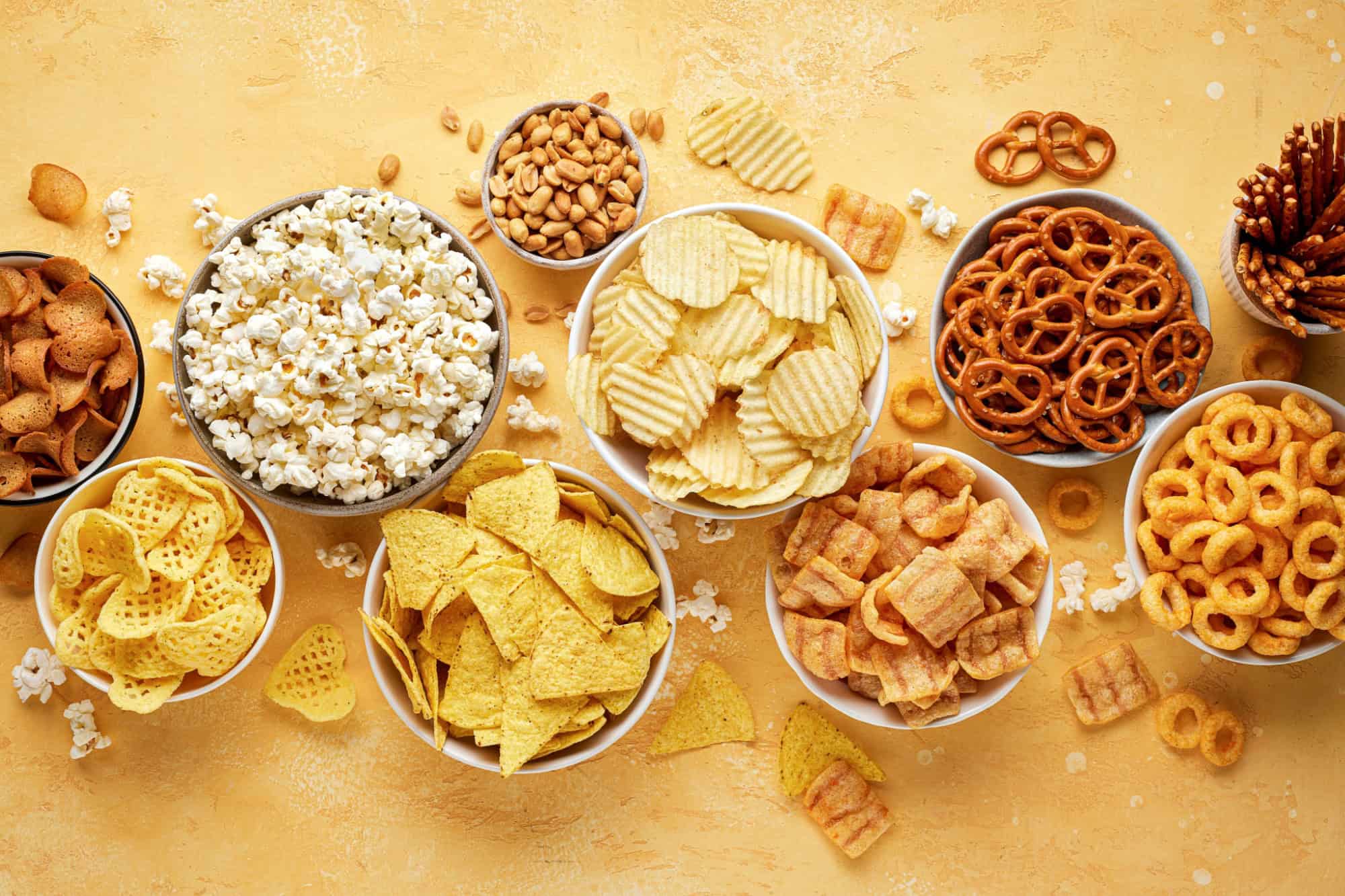 Set of many bowl with different salty snacks on bright yellow background. Chips, popcorn, nuts, nachos and pretzels. Party food. Top view