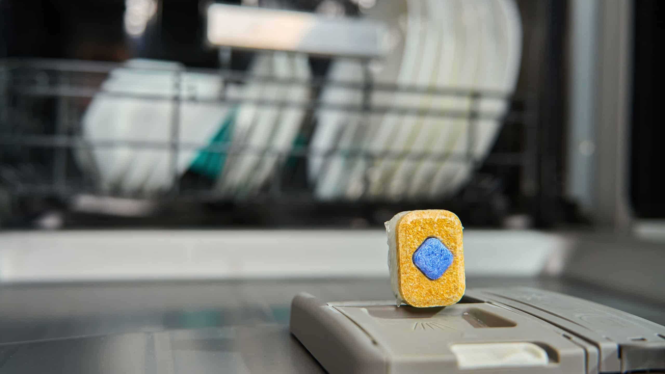 Close-up of dishwasher detergent tablet placed on dispenser lid with clean dishes visible in background. Modern dishwashing solution for effective and eco-friendly cleaning