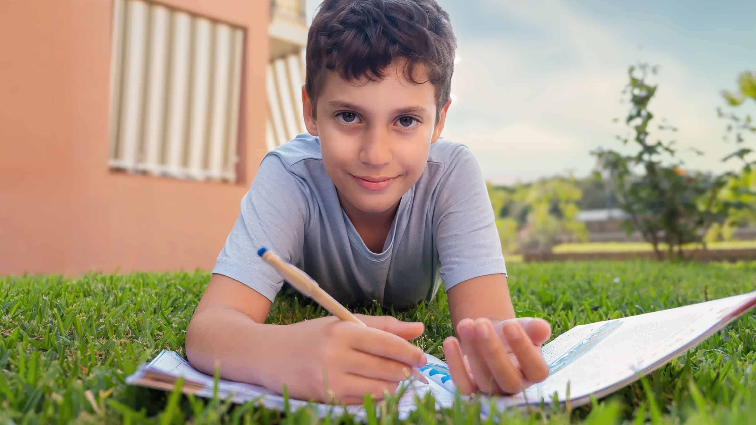 A cheerful young boy studying outdoors, lying on the grass with a notebook and pen. A relaxed and engaging learning moment in nature.