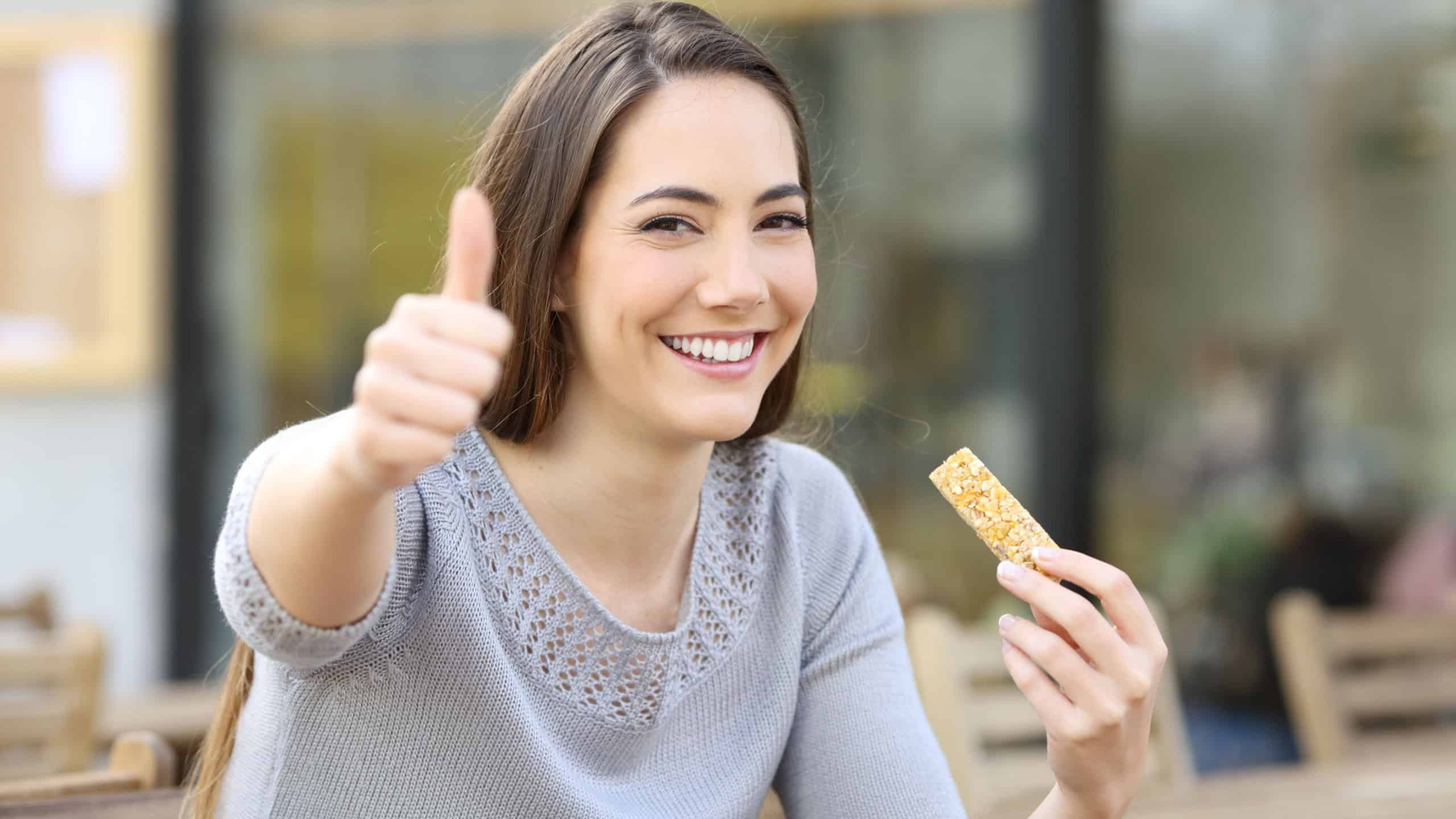 Happy woman eating cereal bar in a restaurant terrace with thumbs up looking at camera
