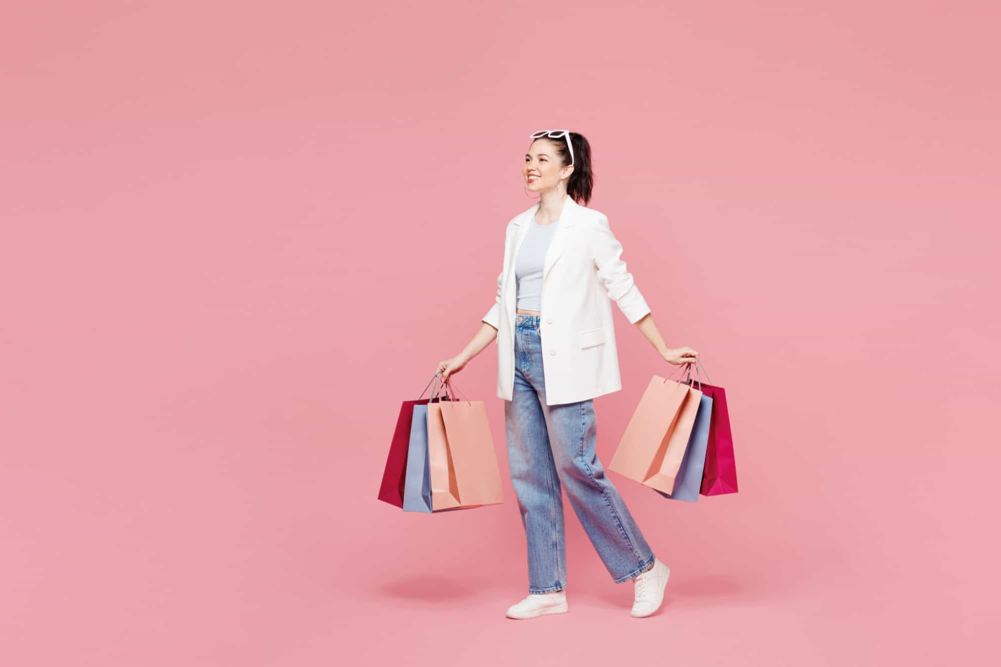 Full body side profile view young smiling woman she wear white clothes hold shopping paper package bags walk go isolated on plain light pastel pink color background. Black Friday sale buy day concept