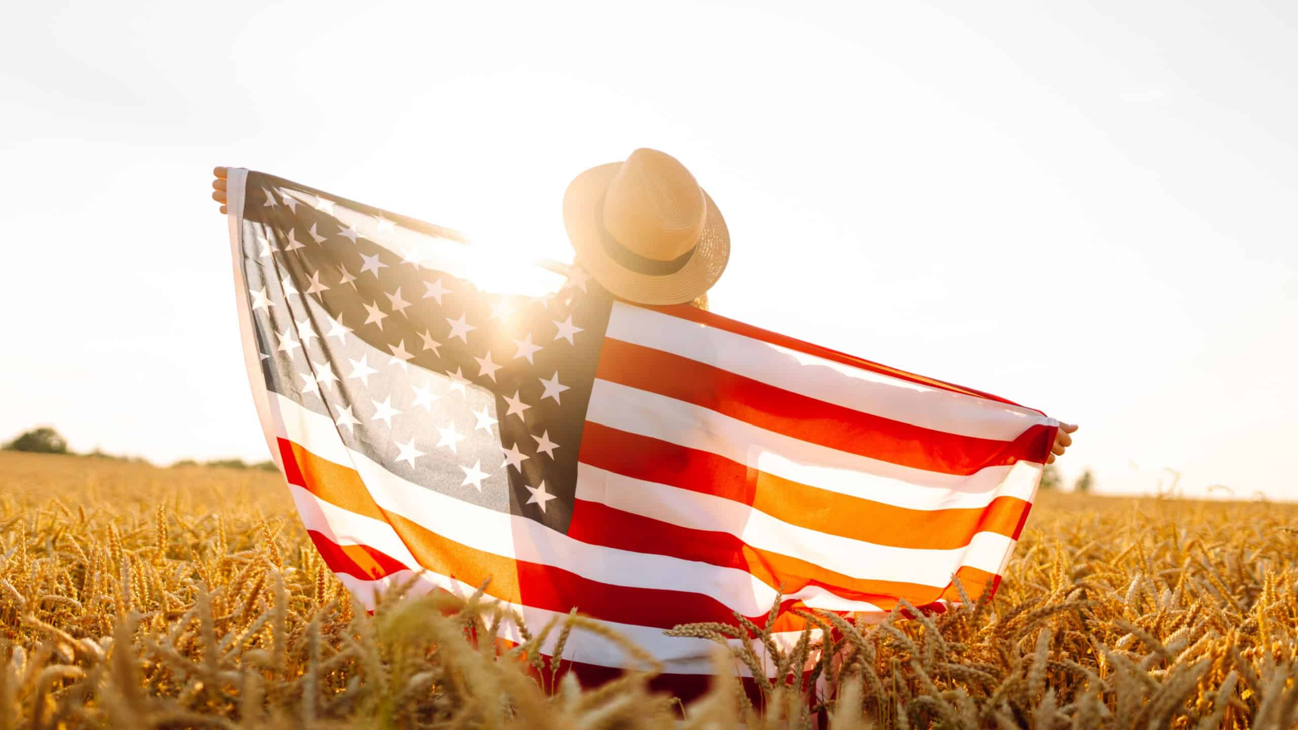 Rear view of happy woman in straw hat holding American flag in sunset agricultural field. Young woman having fun and feeling freedom outdoors. Holiday, fun concept.
