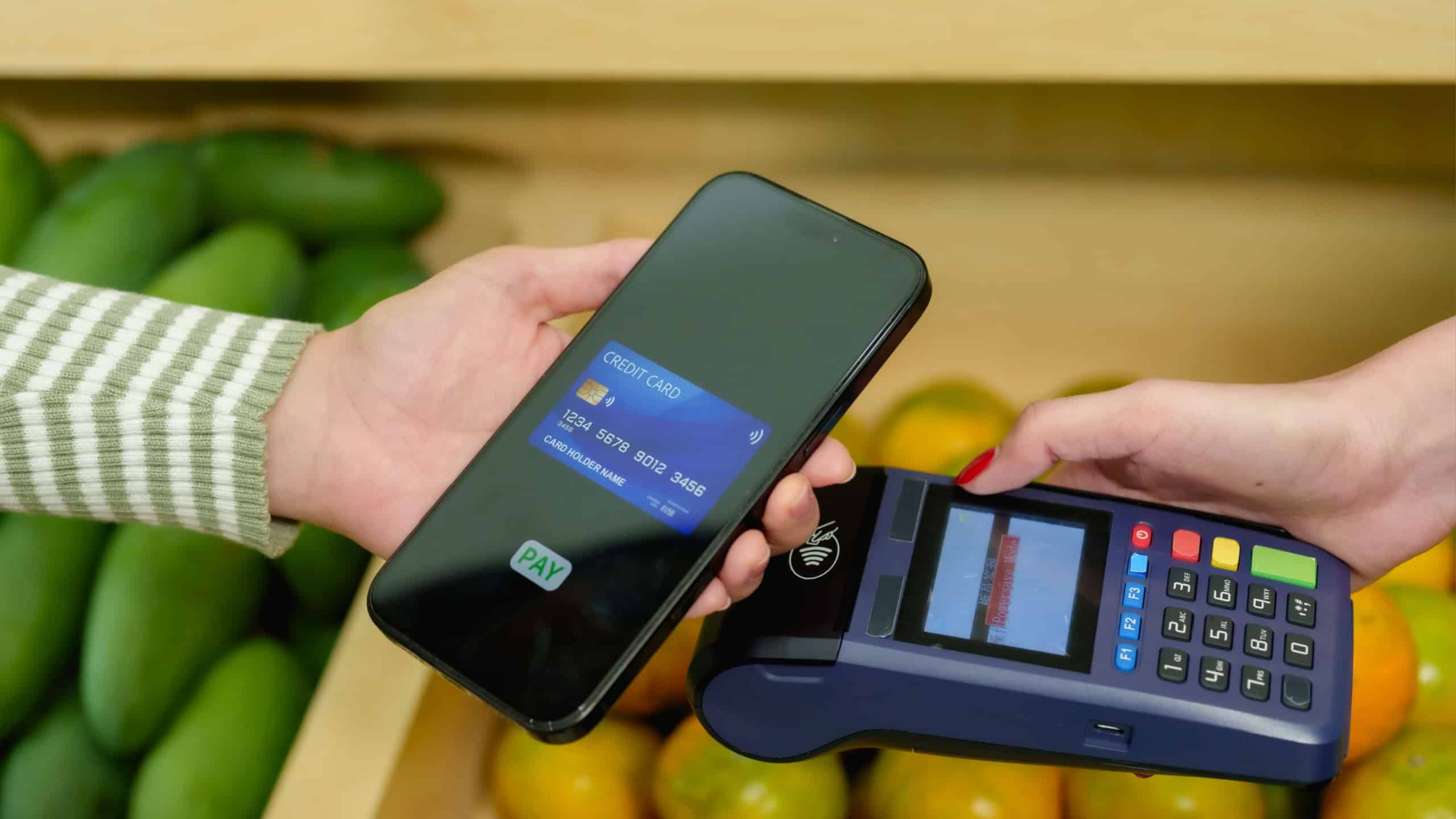 Caucasian adult female using smartphone to tap wireless payment terminal for contactless credit card purchase inside tropical fruit shop operated by Asian female small business shop owner.