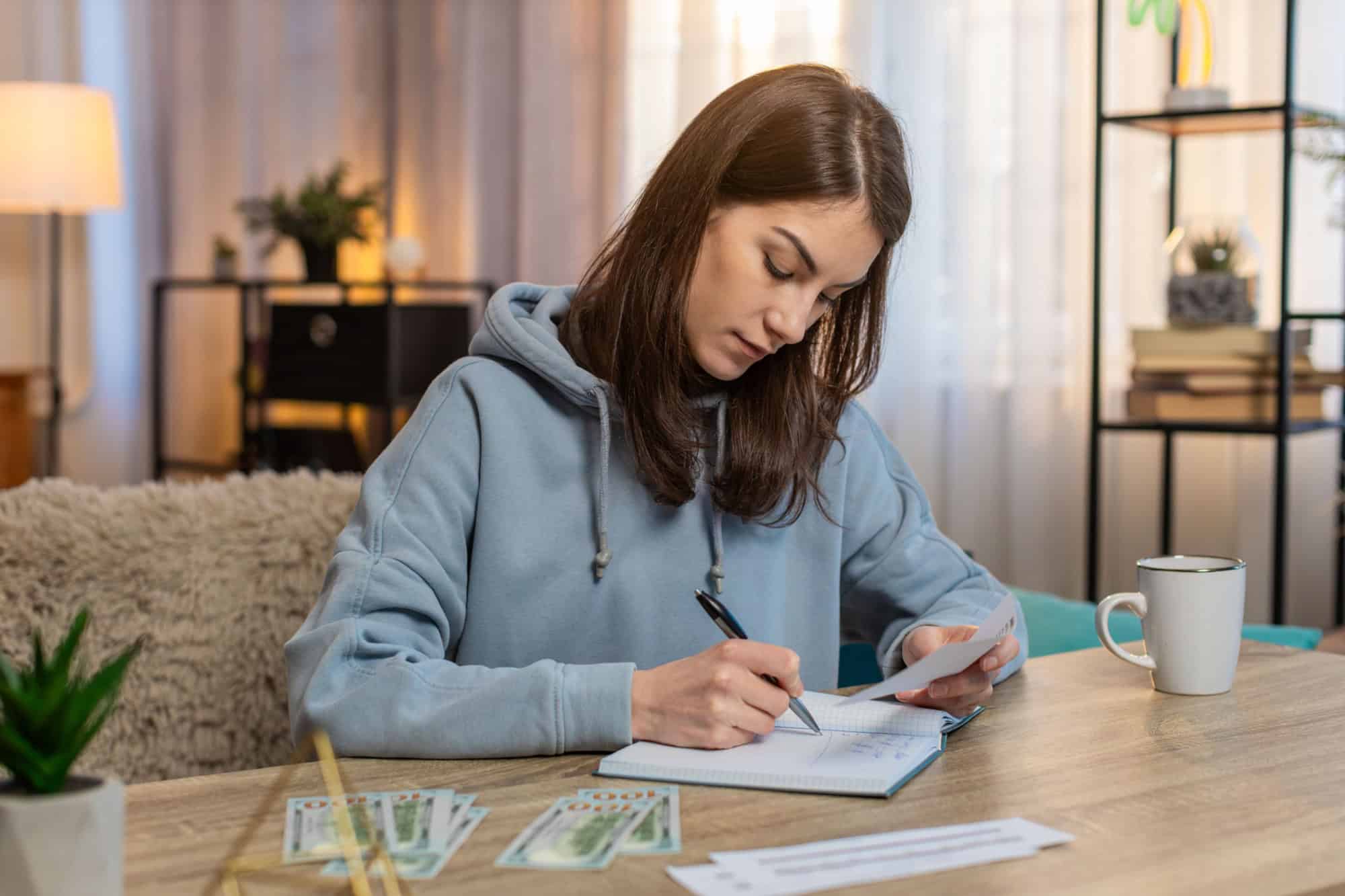 Focused young Caucasian woman making shopping list in notebook sitting at table on sofa looking at receipts and counting money in living room at home. Girl preparing notes for financial cost or budget