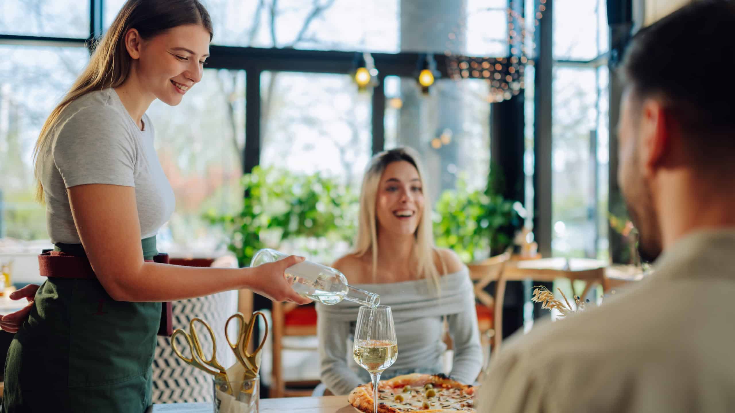 Smiling waitress pouring white wine for happy couple enjoying pizza in modern restaurant, celebrating special occasion