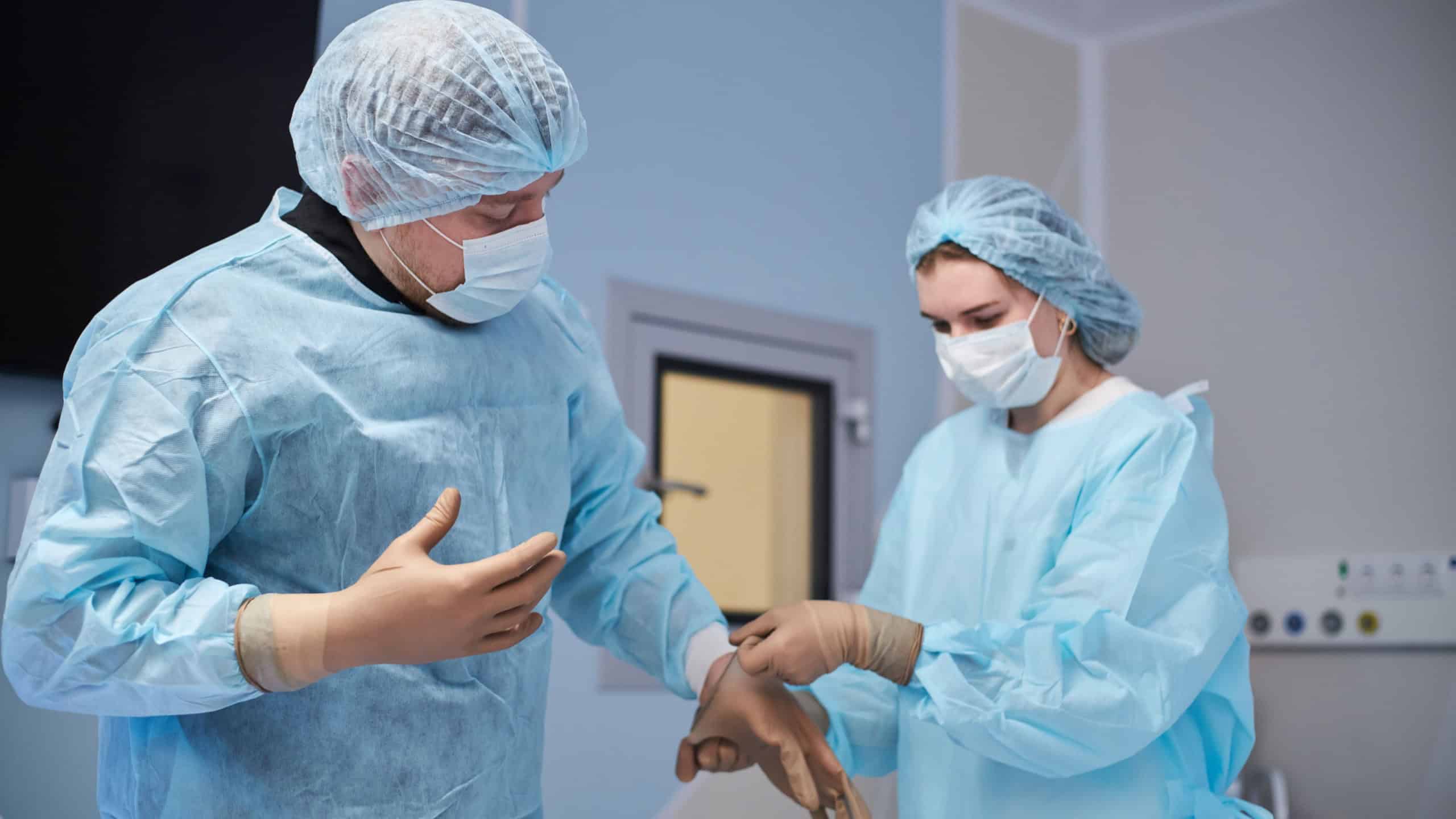 Middle aged man preparing for surgery with woman assisting by putting on sterile gloves in operating room, both wearing surgical masks and gowns