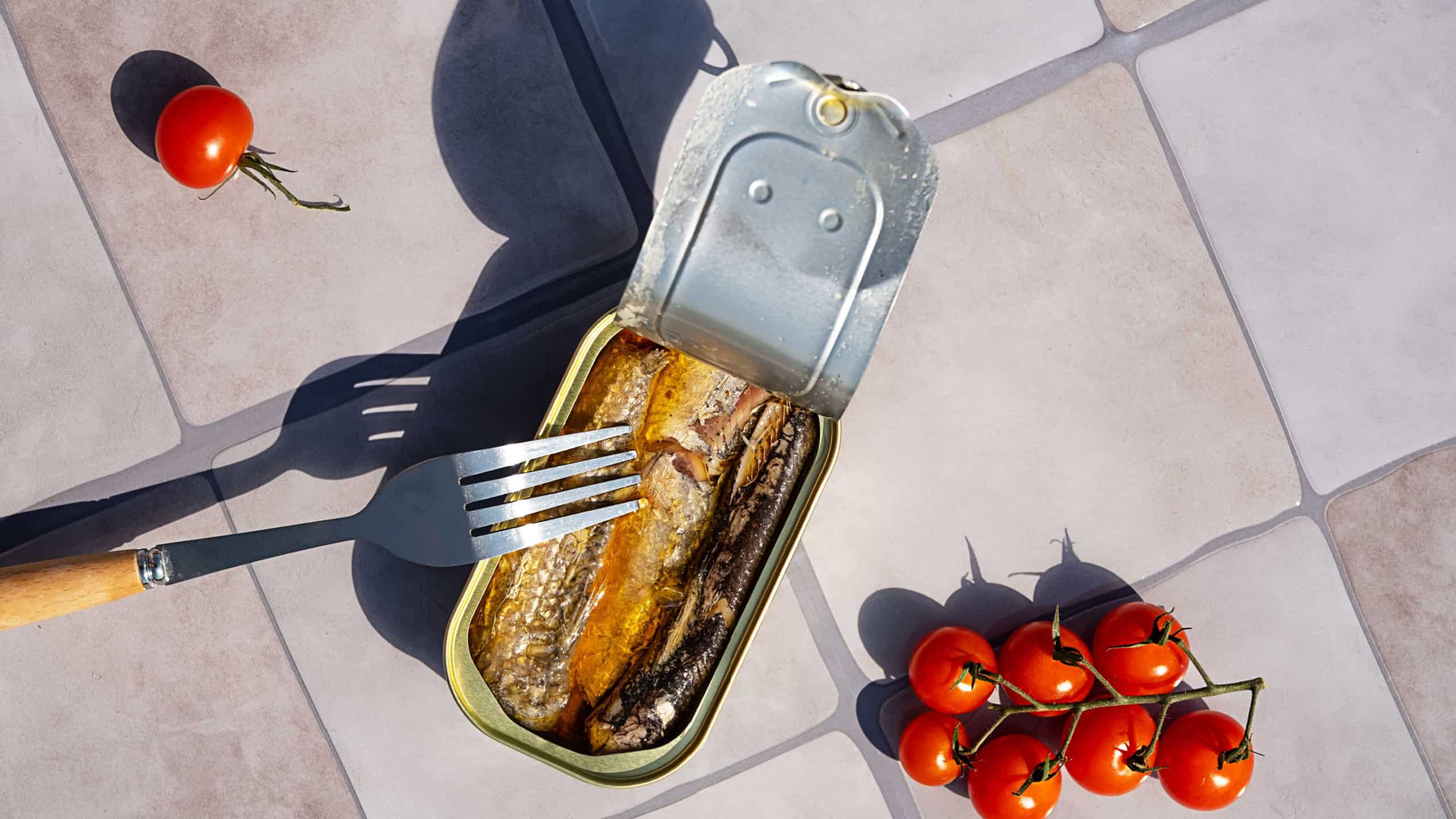 Tin of sardine with fork and tomato on gray tile. Open canned fish for quick dish concept.