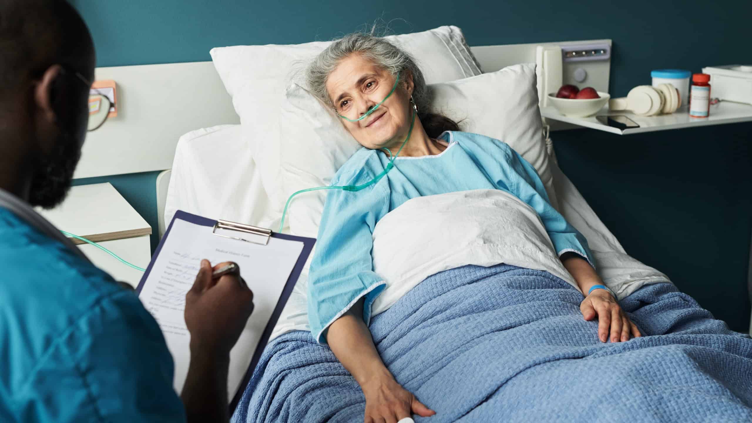 Senior Caucasian woman lying in hospital bed wearing oxygen tube, smiling at Black man healthcare worker taking notes on clipboard, medical monitoring device on finger, clinical setting