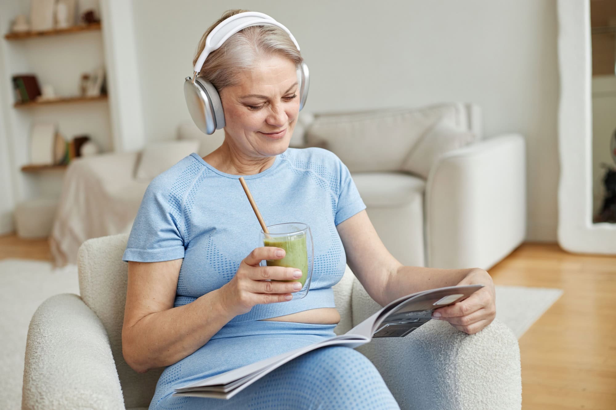 Senior Caucasian woman sitting in chair listening to music with headphones while smiling and holding glass of green smoothie, reading magazine in modern living room