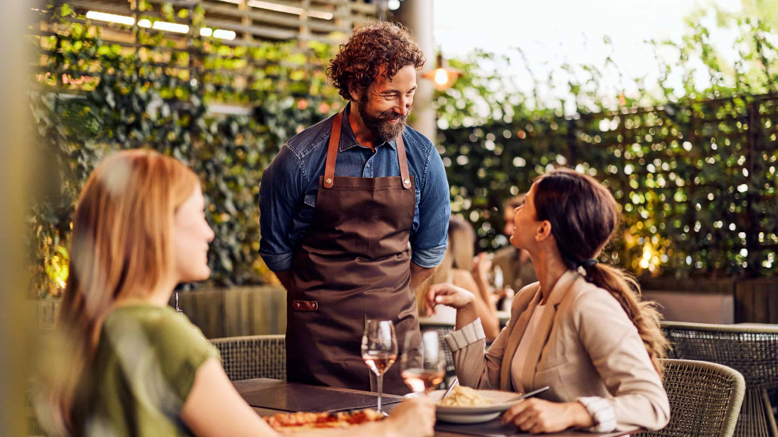 Warm restaurant scene with waiter engaging female diners in outdoor setting.