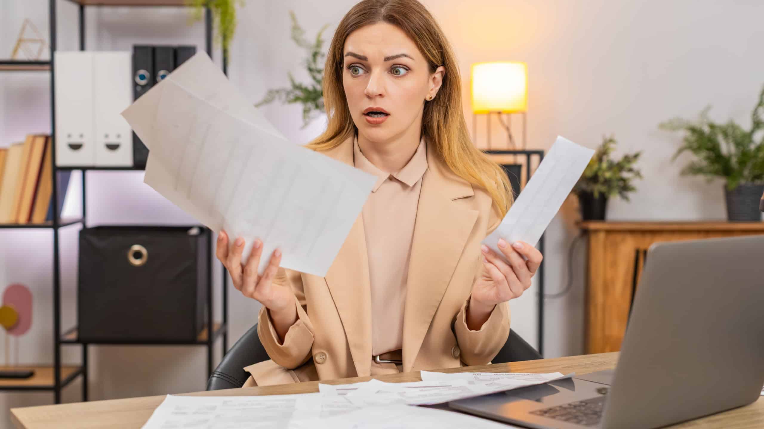 Middle-aged businesswoman at home office looks at bills invoices receipt on laptop sees costs go up frowns confused and unhappy. Freelancer girl at table writes savings plan sets targets prints page